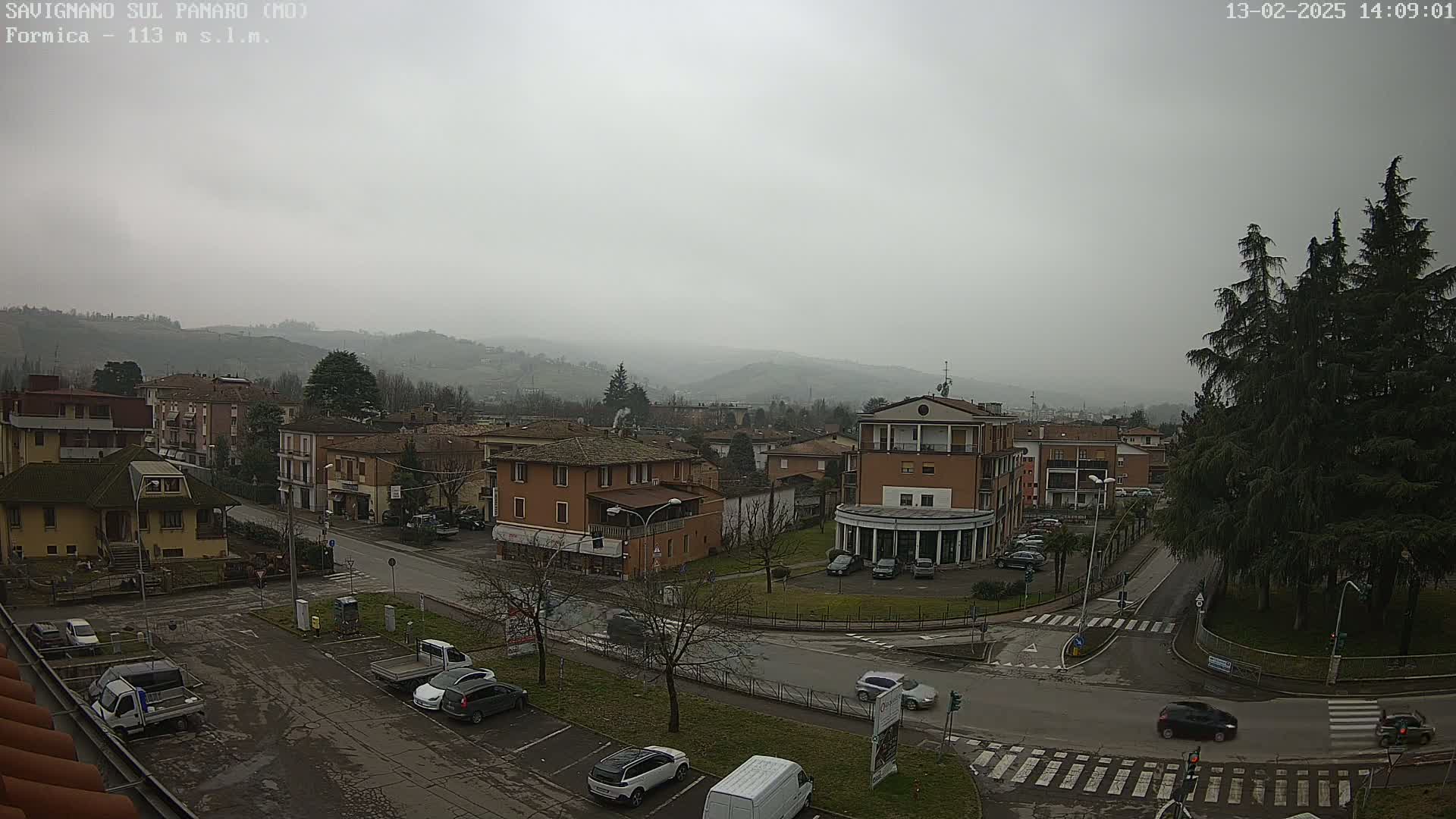 A misty, overcast view of a town nestled in a valley, with several cars parked in a lot and a few driving on the roads.