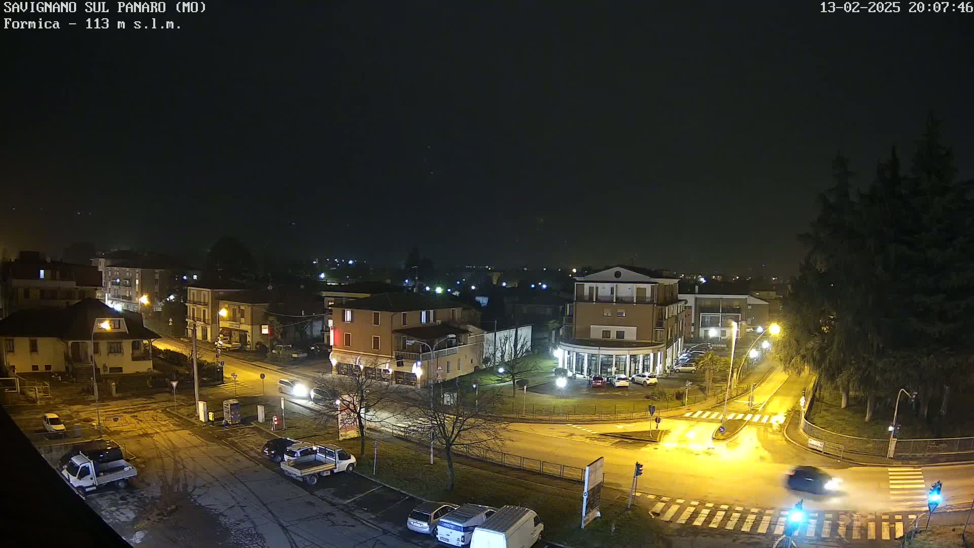 A nighttime, aerial view of a town square with several buildings, vehicles, and streetlights, under a dark, clear sky.