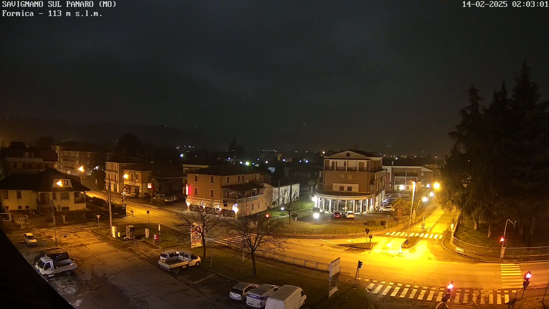 A nighttime view of a town square with several buildings, streetlights illuminating the roads and a few parked vehicles, under a hazy, dark sky.