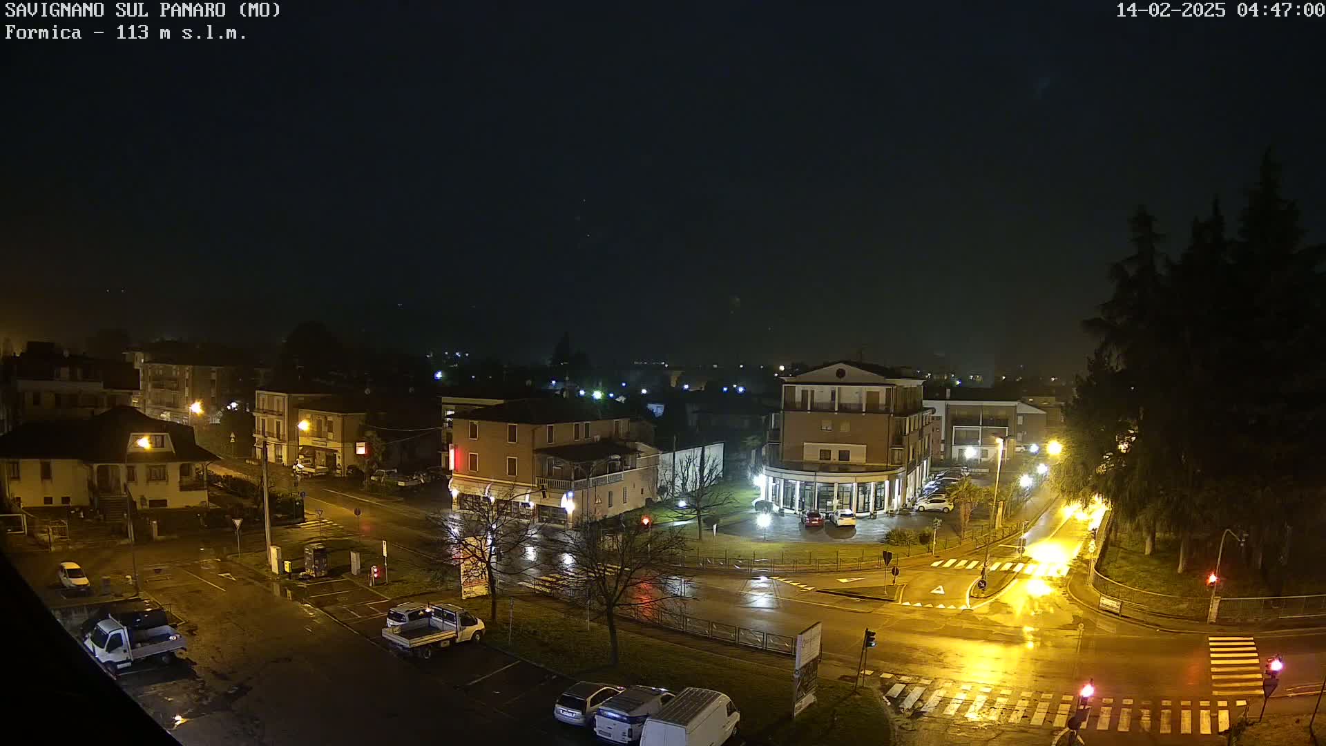 A rain-slicked town square at night, illuminated by streetlights, with several buildings and parked vehicles visible.