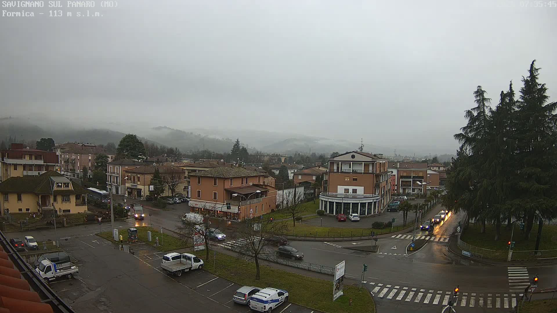 A misty, overcast day reveals a town square with light traffic, surrounded by low-rise buildings and hills shrouded in fog.