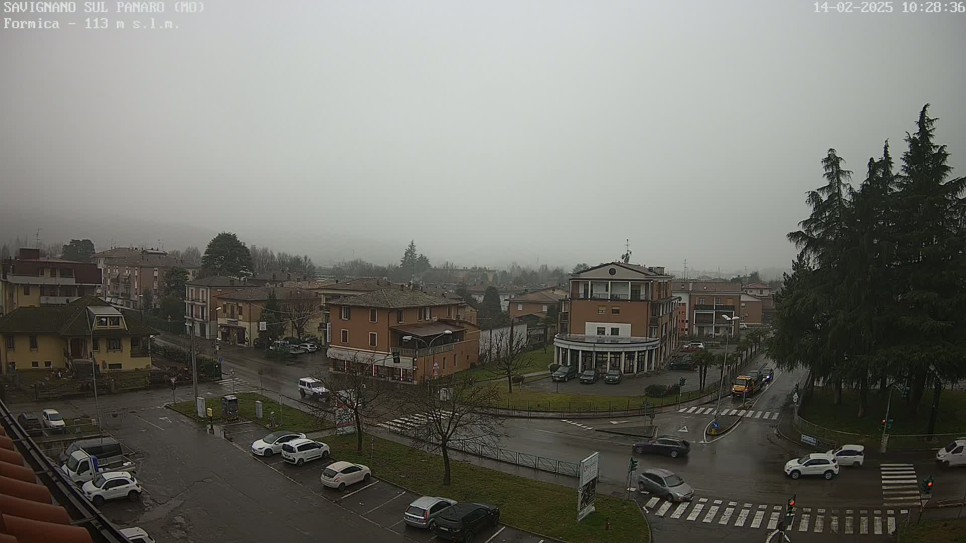A wet, overcast town square is viewed from above, showing several cars parked and driving on a wet street, surrounded by low-rise buildings and evergreen trees.