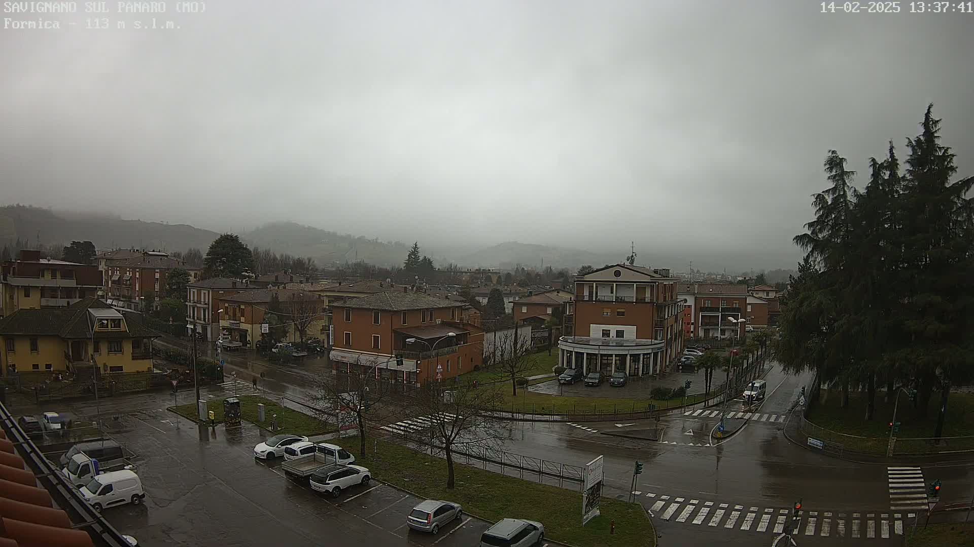 A rain-soaked town square with cars parked and driving in the wet streets, surrounded by buildings and hills under a gray, overcast sky.