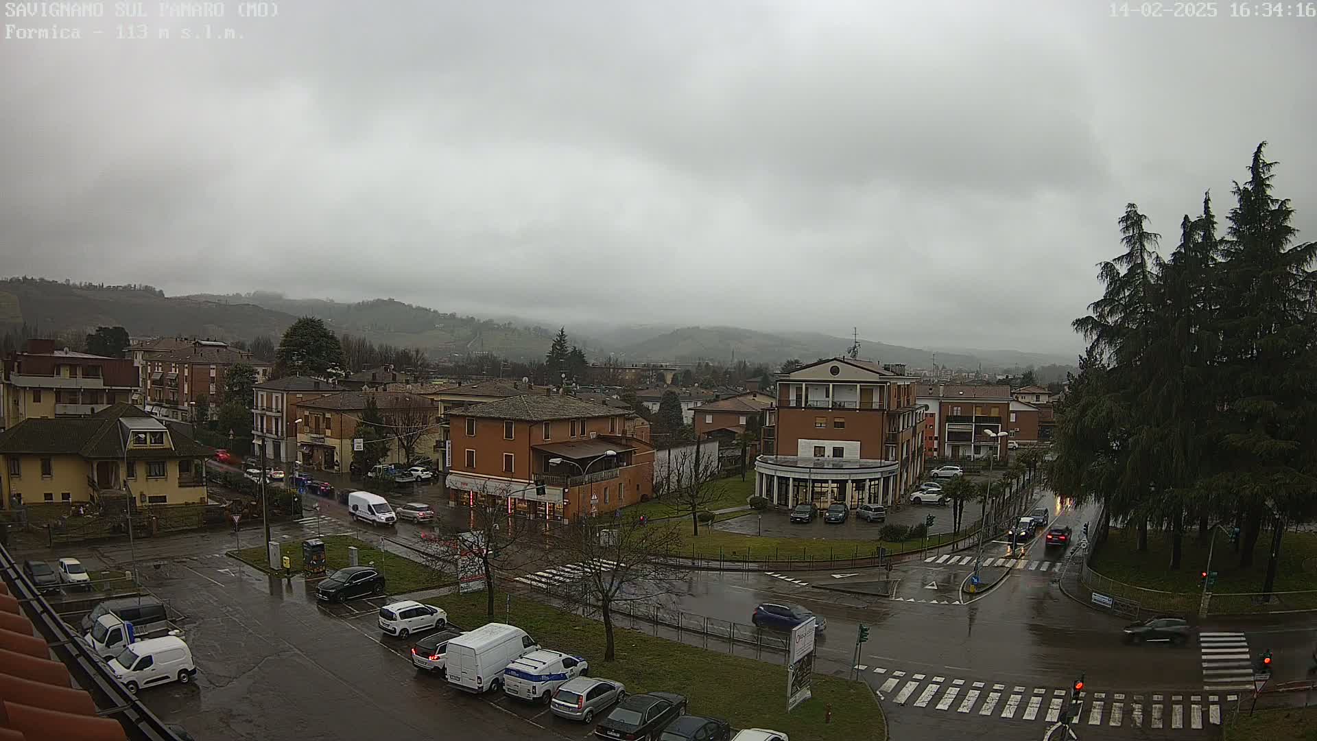 A rain-soaked town square with several cars parked and driving around it, surrounded by low-rise buildings and hills under a cloudy sky.