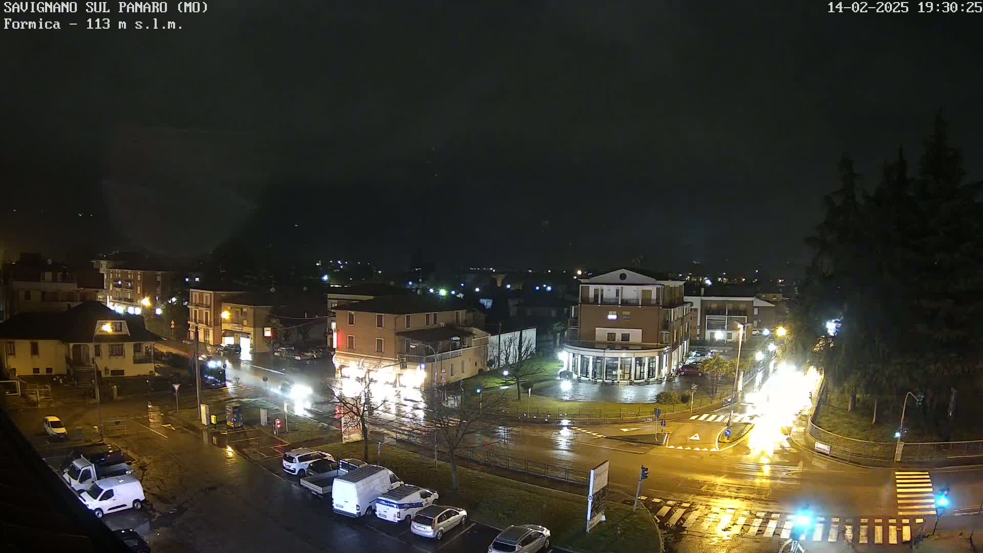 A nighttime view of a town square with wet roads and buildings illuminated by streetlights under a dark, cloudy sky.