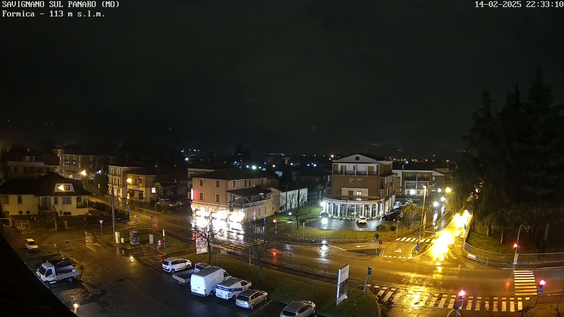 A nighttime, rain-slicked town square with several parked cars, streetlights, and buildings is illuminated by bright yellow streetlights and an area of bright orange light further down the street.