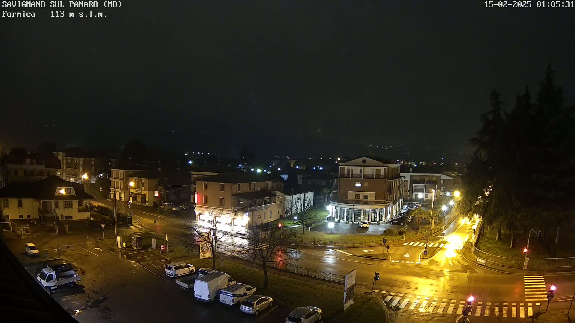 A nighttime view of a town square with wet streets and parked cars under a dark, overcast sky.