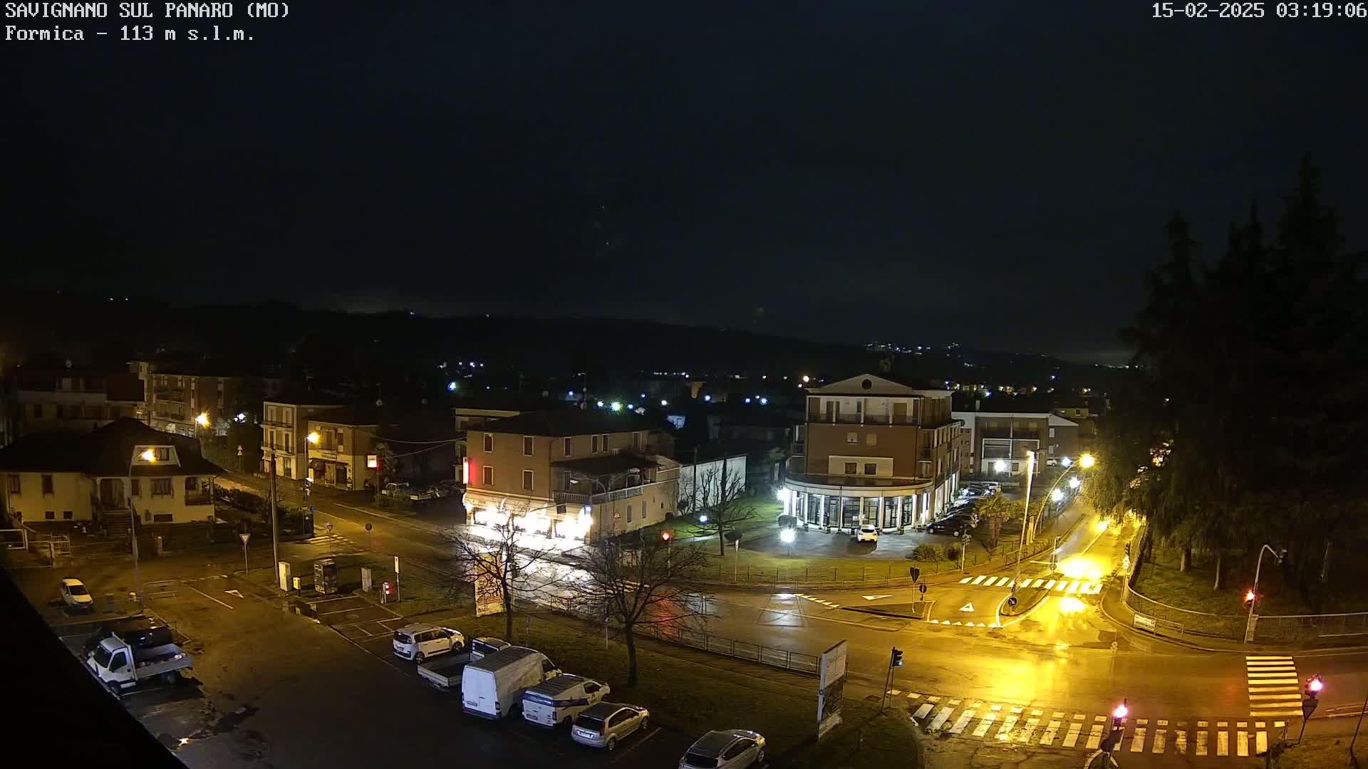 A nighttime, wet street scene shows several buildings and cars parked along the side of a road, illuminated by streetlights.