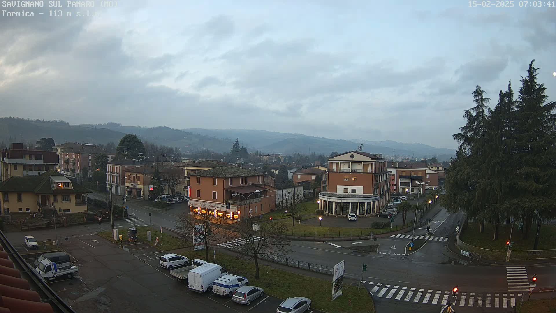 A view of a town square and surrounding buildings under an overcast sky, with some cars parked and a few driving on wet roads.