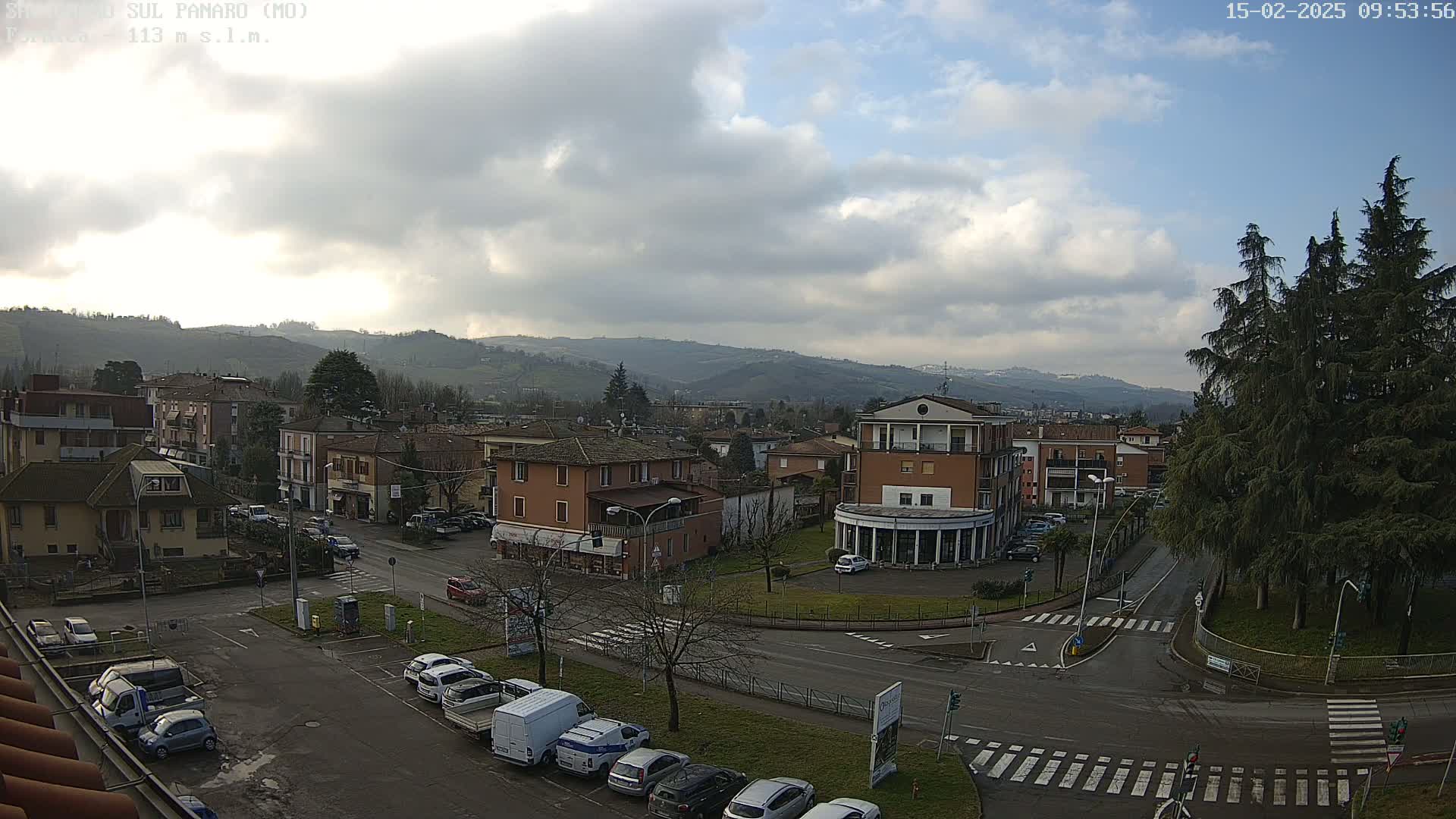 A partly cloudy day reveals a small town nestled in a valley, surrounded by hills, with cars parked in a lot and along the streets.