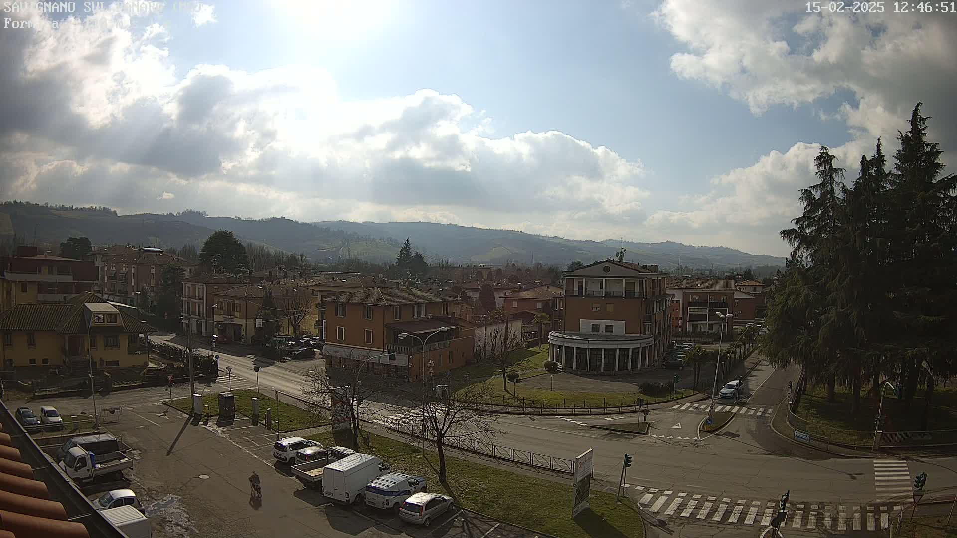 A partly sunny day reveals a town nestled in a valley, surrounded by hills, with vehicles parked in a street and a cyclist riding along a road.