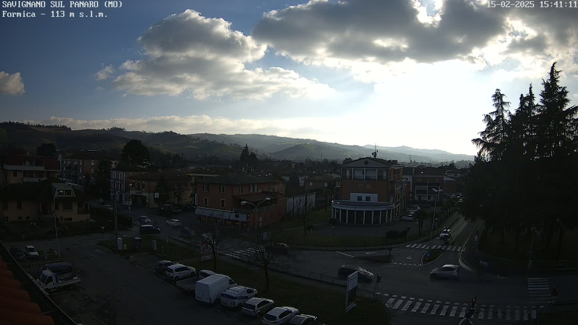 A partly cloudy daytime view of a town nestled in a valley, with cars parked and driving on roads that intersect in a roundabout.