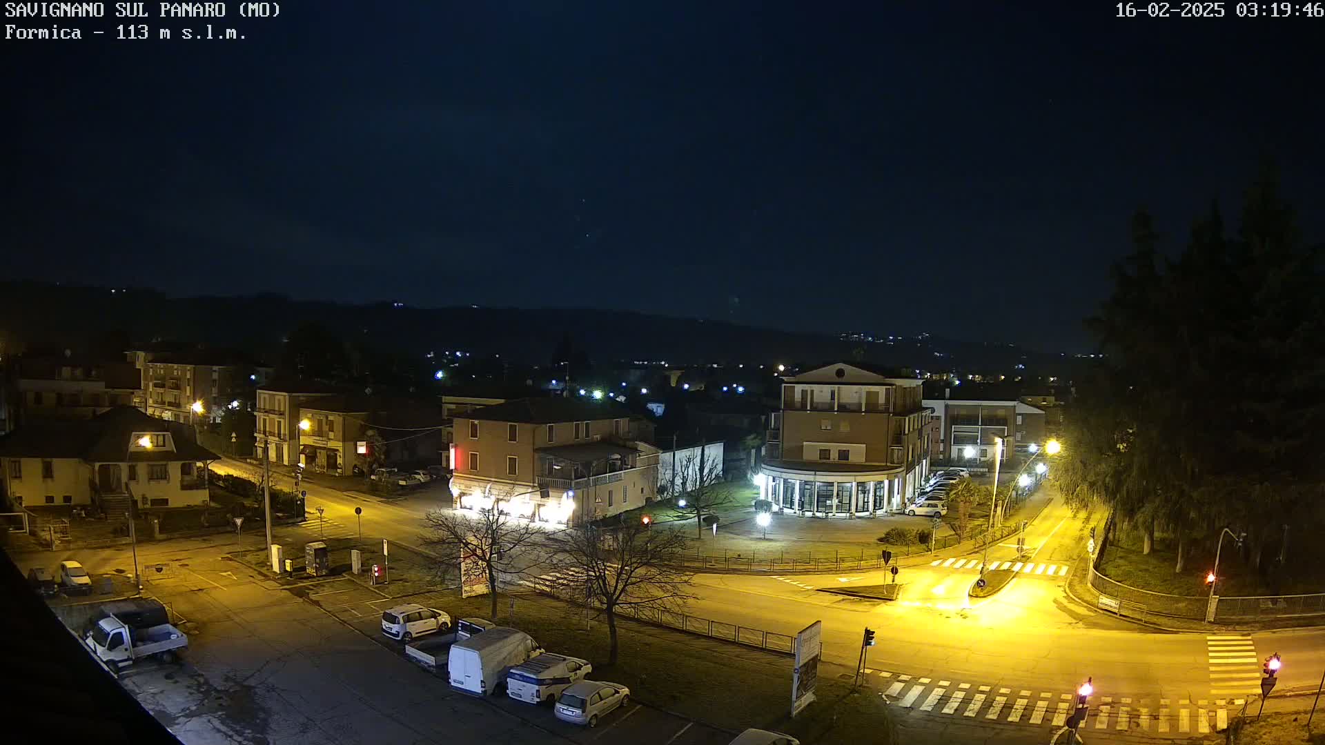 A nighttime aerial view of a town square with parked cars and illuminated buildings under a clear, dark sky.
