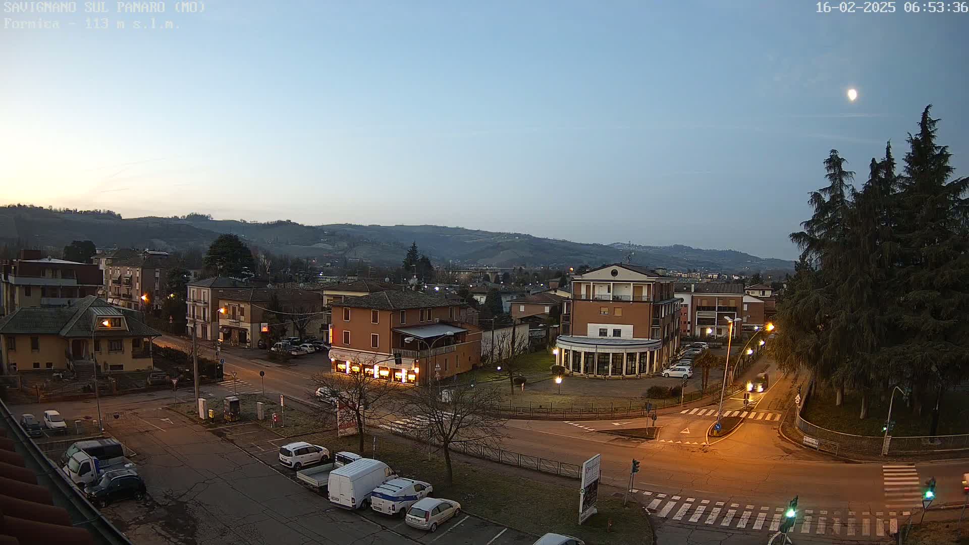A moonlit, pre-dawn view of a town square with several buildings, cars in parking lots, and a roadway intersection, all situated in front of a range of low hills.