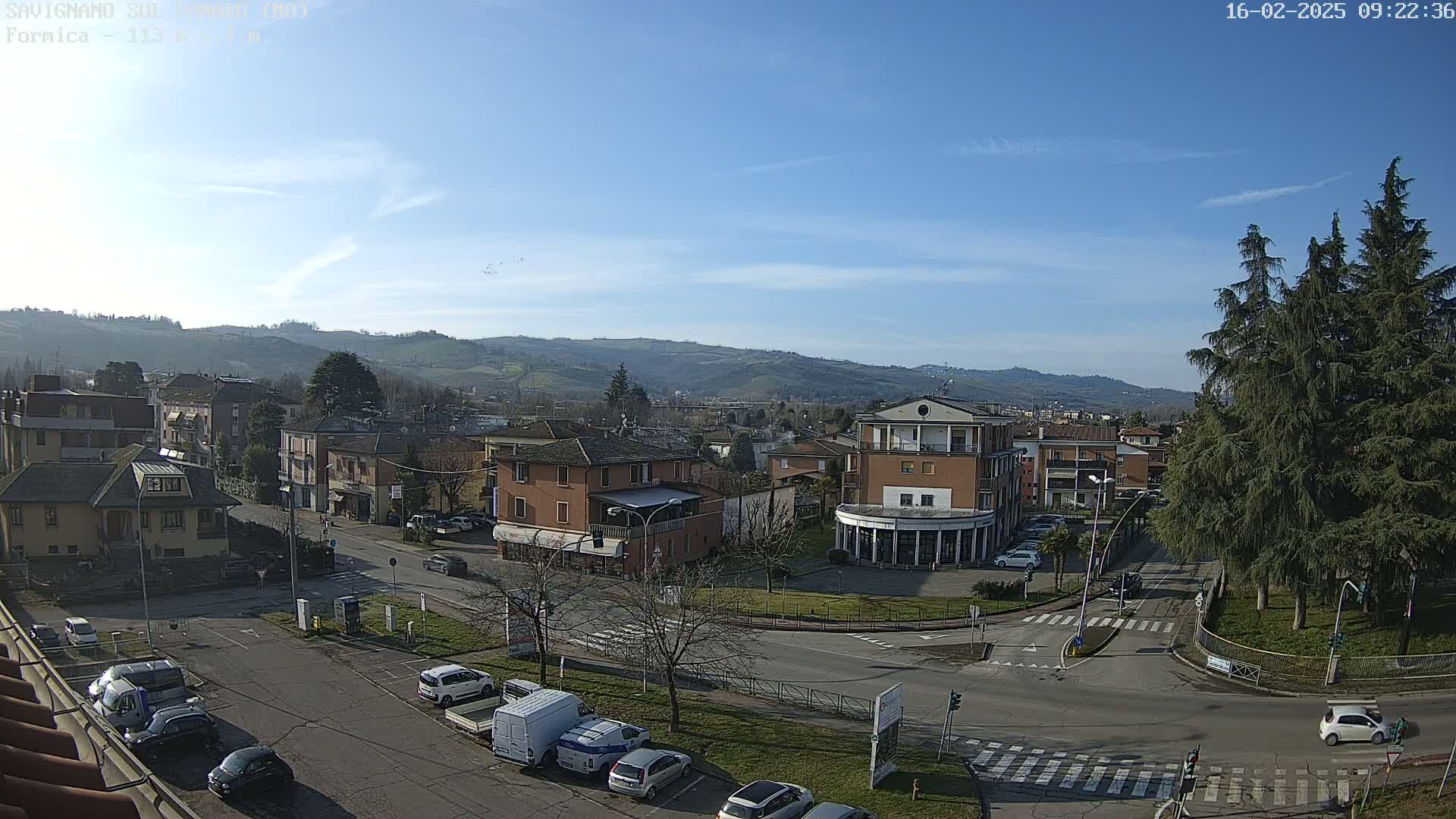 A sunny day reveals a small town nestled in a valley, with cars parked along roads and buildings surrounding a central roundabout.