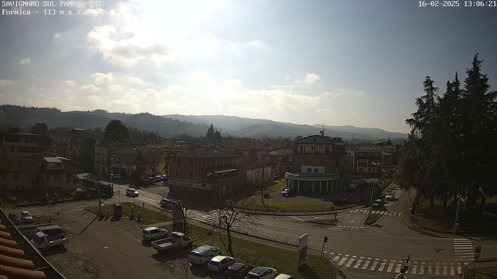 A mostly sunny day reveals a view of a town nestled in a valley, with cars parked in a lot and driving on roads, and hills visible in the distance.