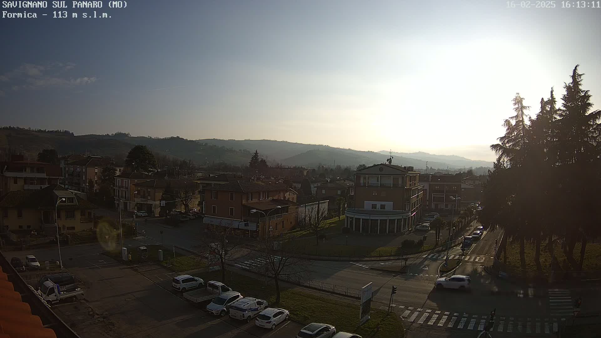 A sunny, clear day overlooks a town nestled in a valley, with cars parked along the roads and several buildings visible, including a large, light-brown building near the center.