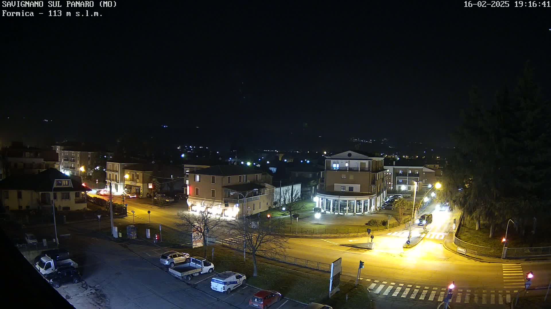A nighttime aerial view of a town square with cars parked and driving on illuminated streets under a clear, dark sky.