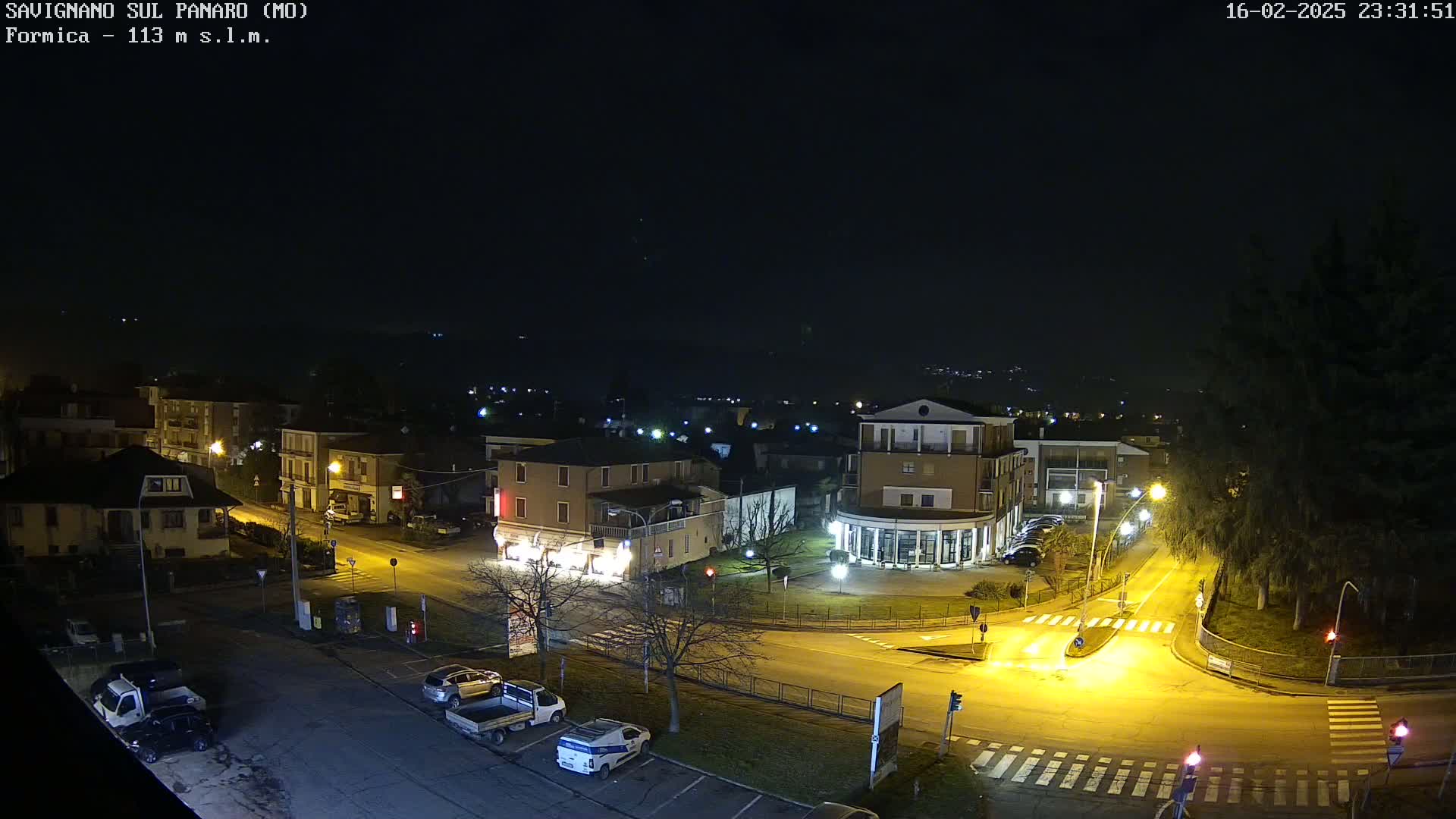 A nighttime aerial view of a town square with several buildings and parked cars, illuminated by streetlights under a clear, dark sky.
