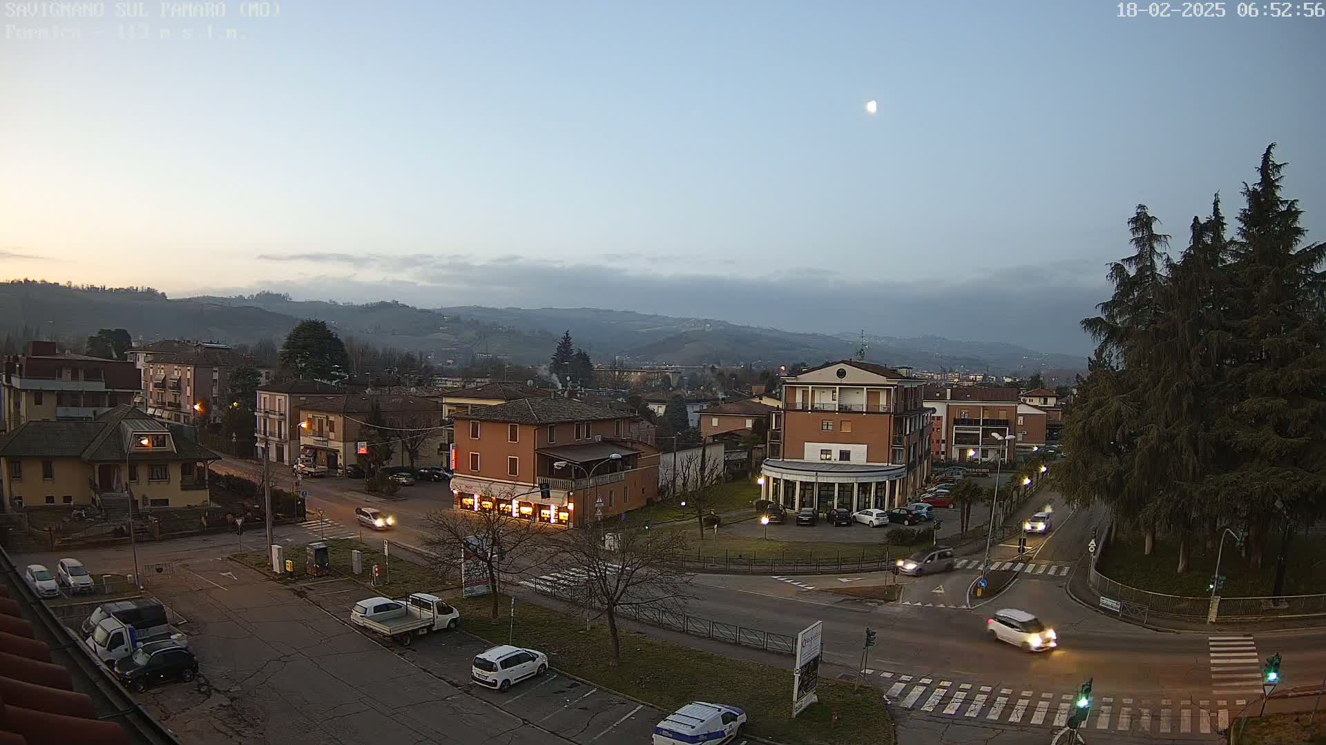 A high-angle, twilight view of a town nestled in a valley, with a few cars on the roads and a crescent moon visible in the pale sky.