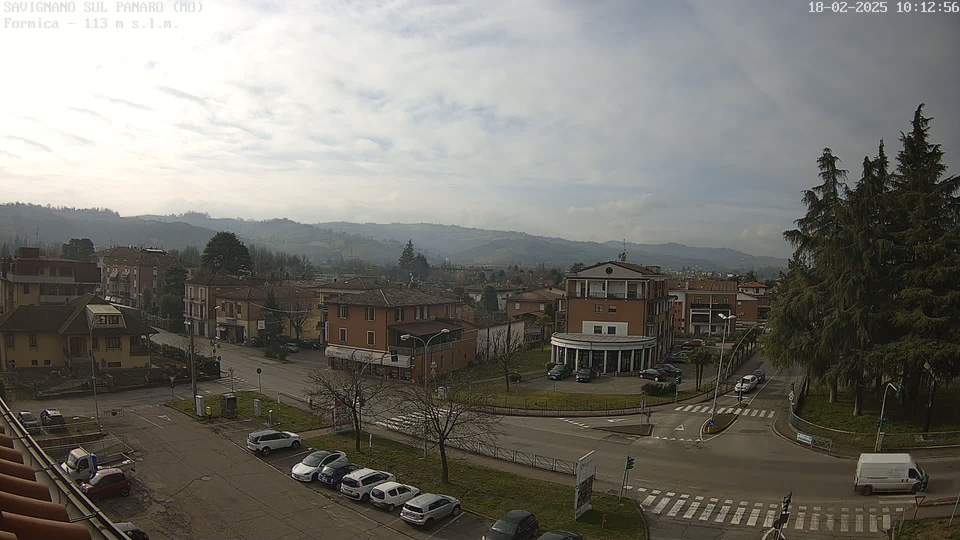 A mostly sunny view of a town square in a valley, surrounded by low hills and evergreen trees, with several cars parked along the streets.