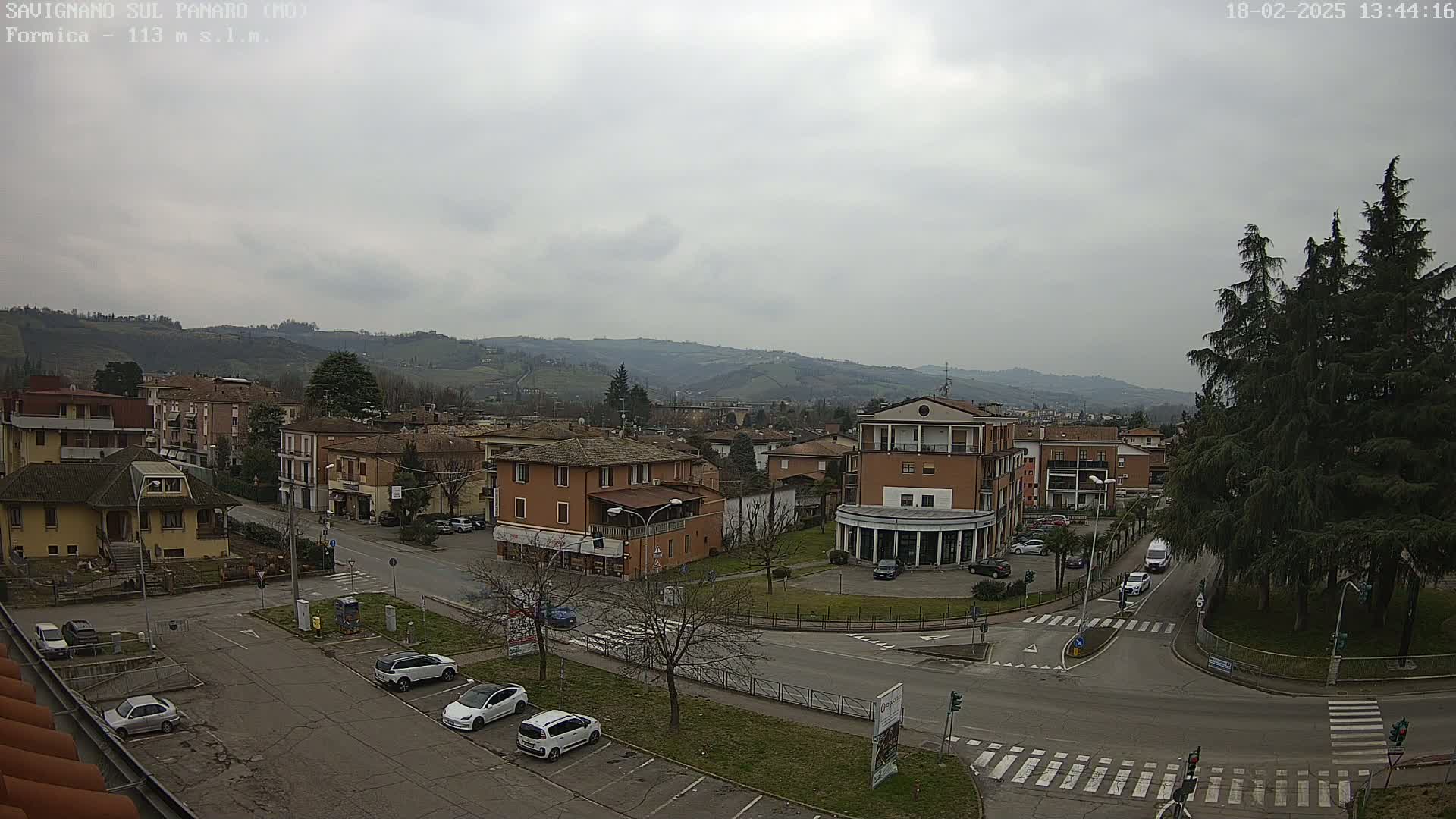 An overcast day reveals a town nestled in a valley, with cars parked in a square surrounded by buildings, and hills visible in the background.