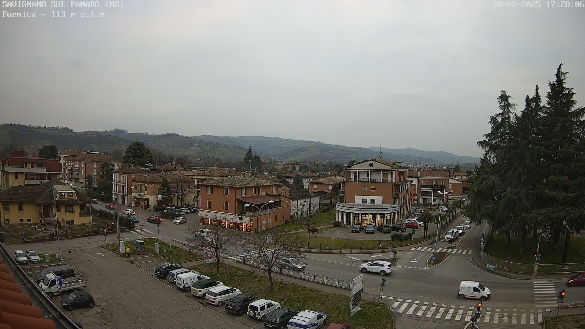 An overcast day reveals a town square surrounded by low-rise buildings and parked cars, with hills visible in the background.