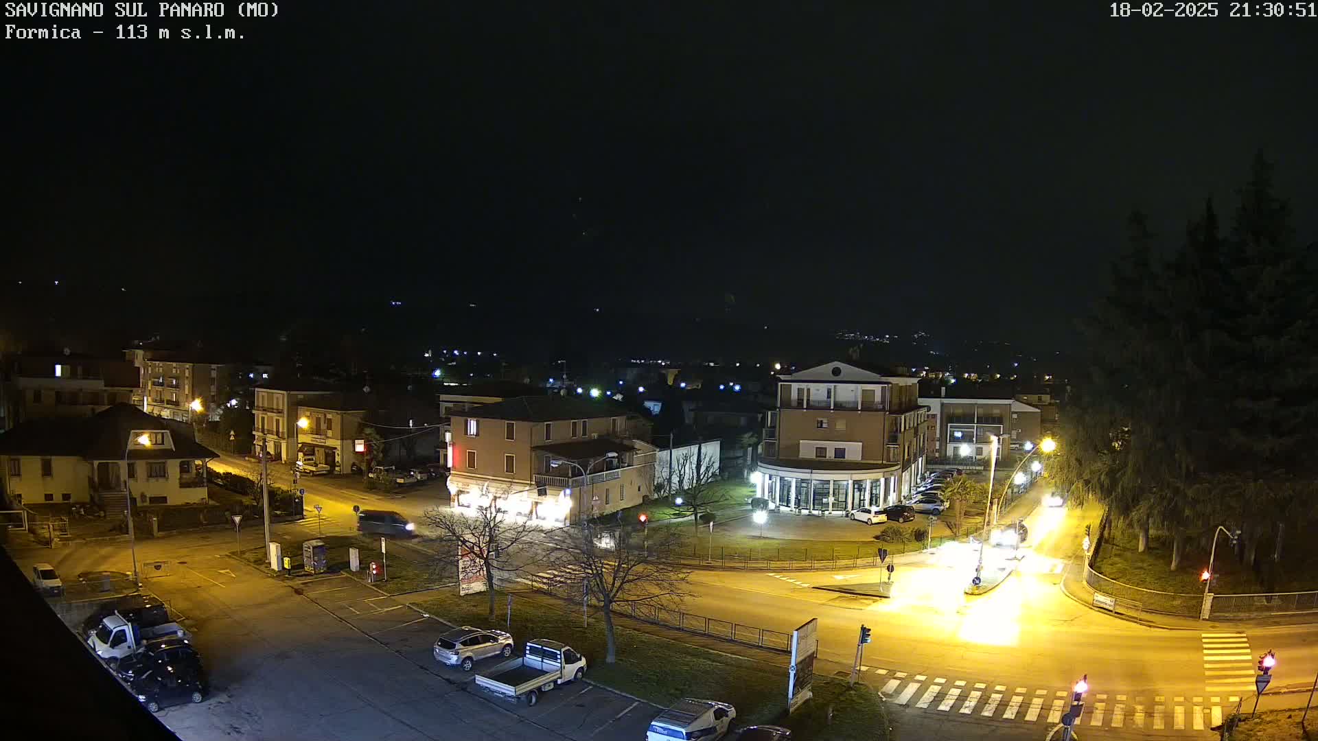 A nighttime aerial view of a town square with cars and buildings illuminated by streetlights under a clear, dark sky.