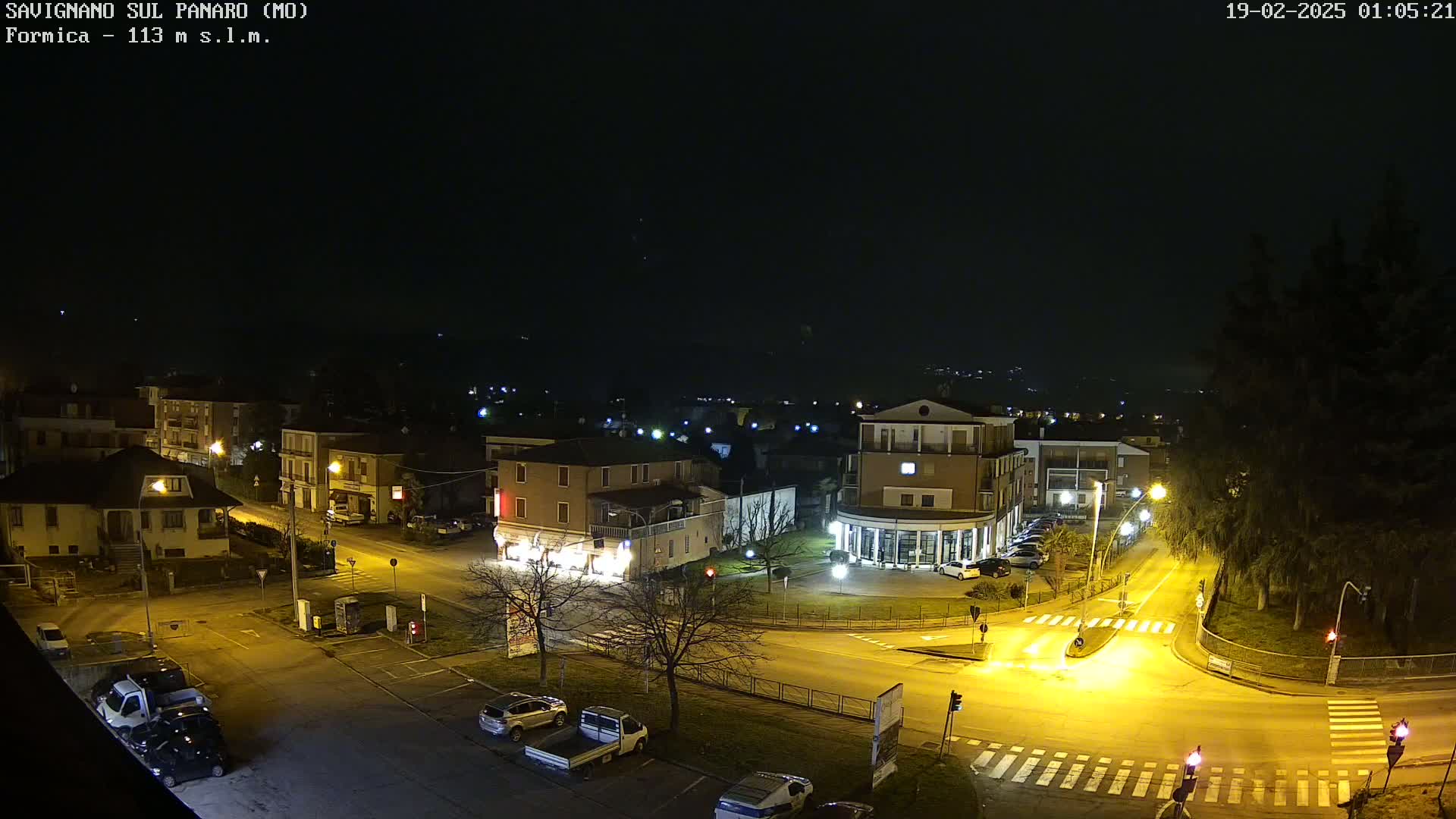 A nighttime aerial view of a town square with several buildings, cars, and streetlights illuminated under a clear, dark sky.