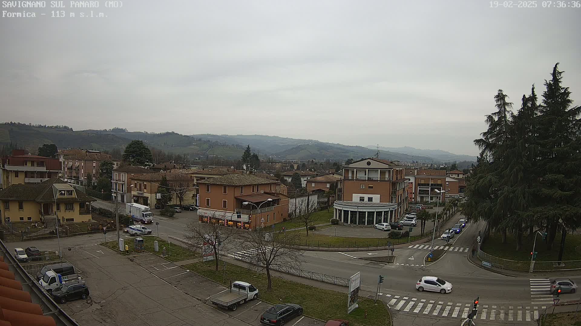 A view of a town under an overcast sky, showing low-lying buildings and roads with several cars parked and driving, all situated in a valley surrounded by hills.