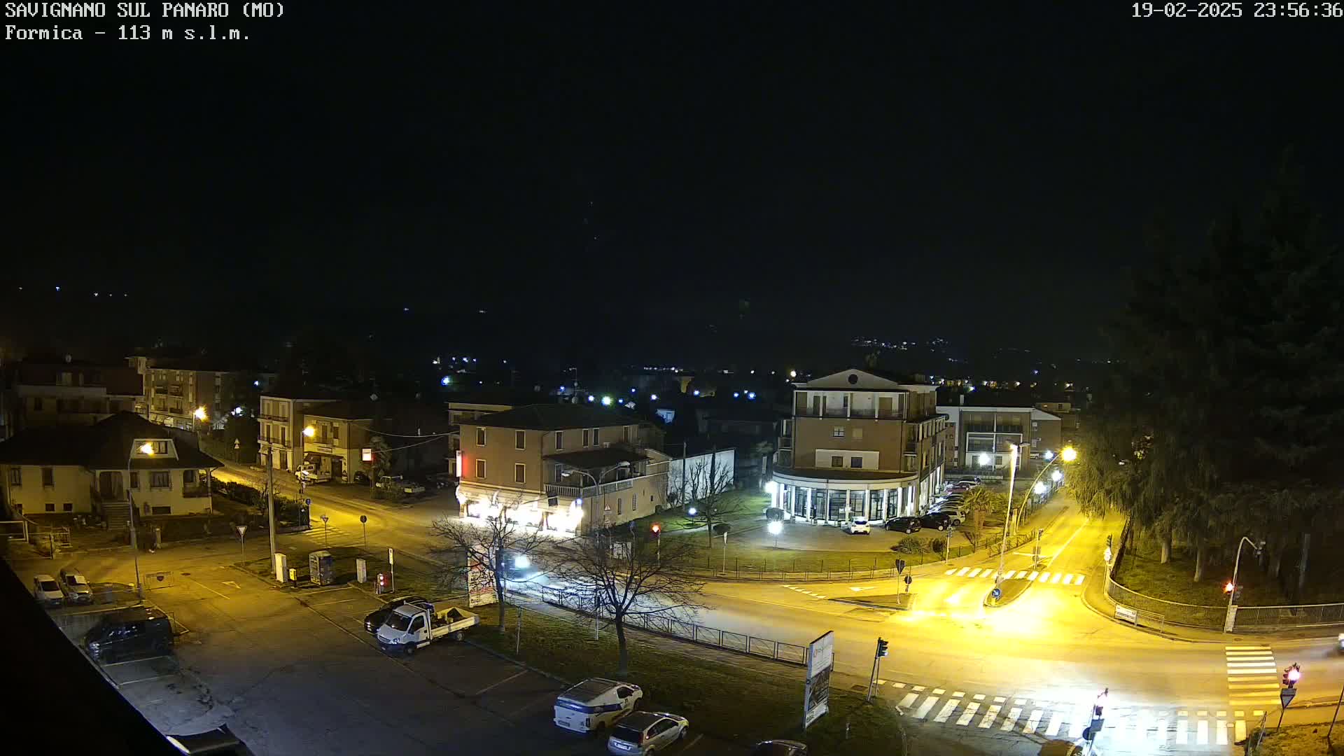 A nighttime aerial view of a town square with buildings and vehicles, under a clear, dark sky.