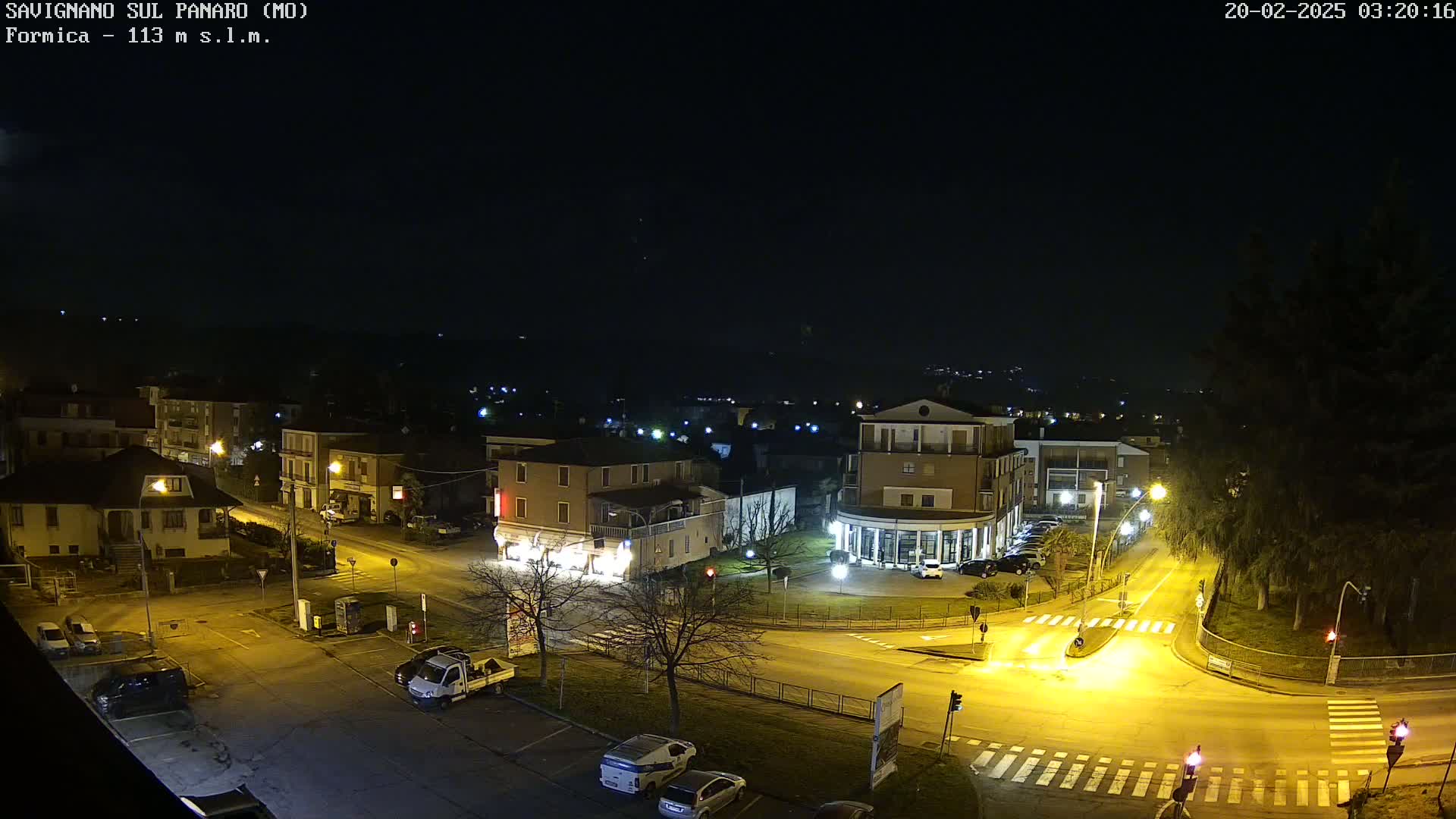 A nighttime, overhead view of a town's intersection with cars parked and driving, streetlights illuminating the scene under a clear, dark sky.