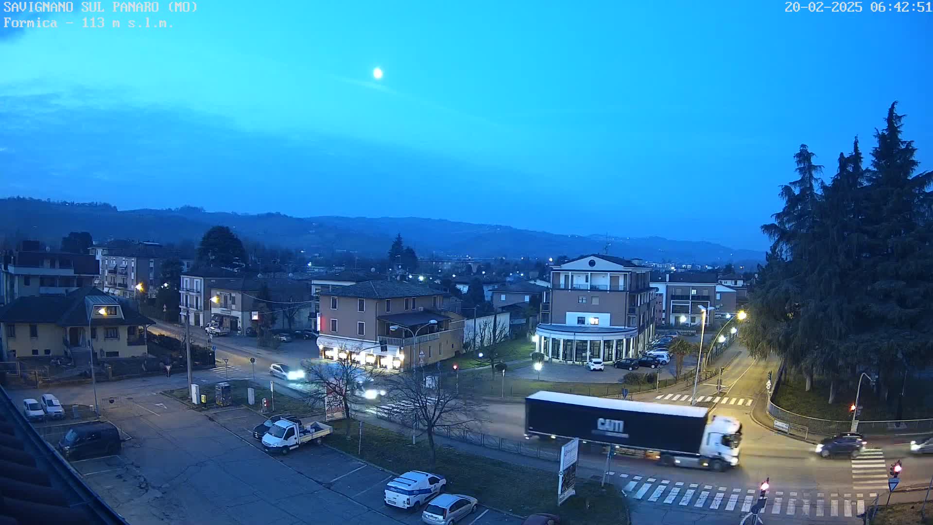 A twilight view of a town square with several buildings, vehicles, and trees under a clear, blue-tinged sky, illuminated by streetlights.