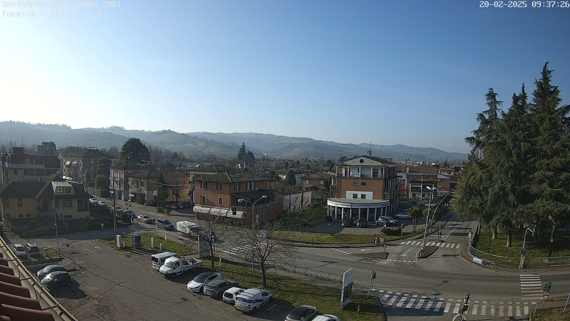 A sunny day overlooking a town square with several buildings, cars, and trees, and hills in the background.