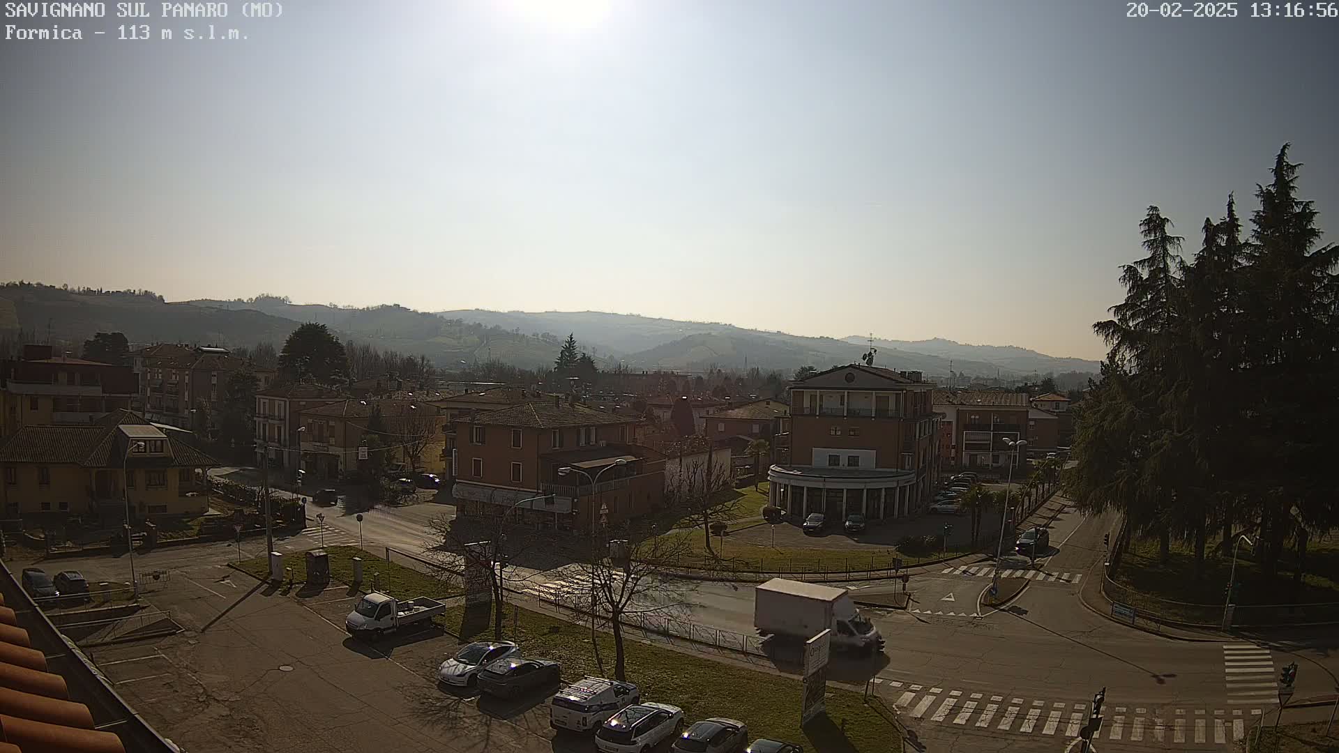 A sunny day reveals a town nestled in a valley, with cars parked in a lot and several buildings visible, surrounded by hills and evergreen trees.
