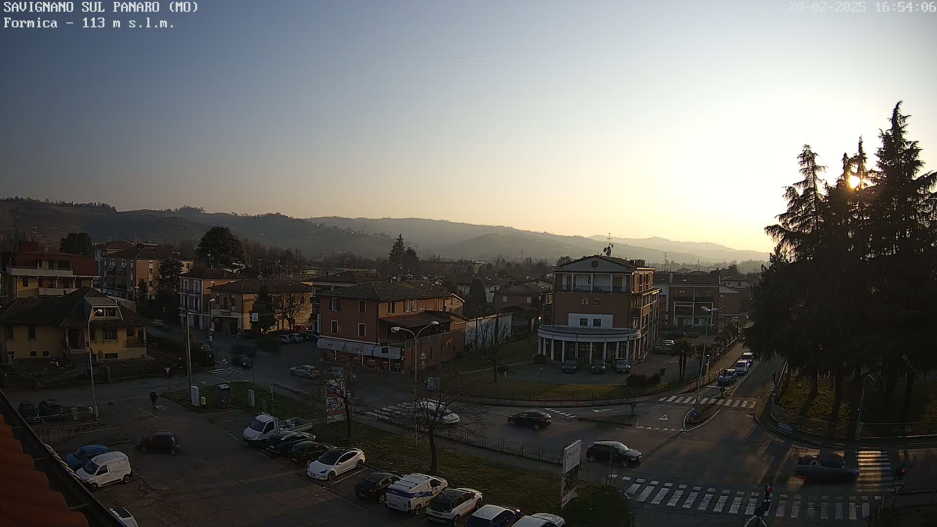 A sunlit town square with cars parked and driving around, surrounded by low buildings and hills in the background under a clear sky at sunset.