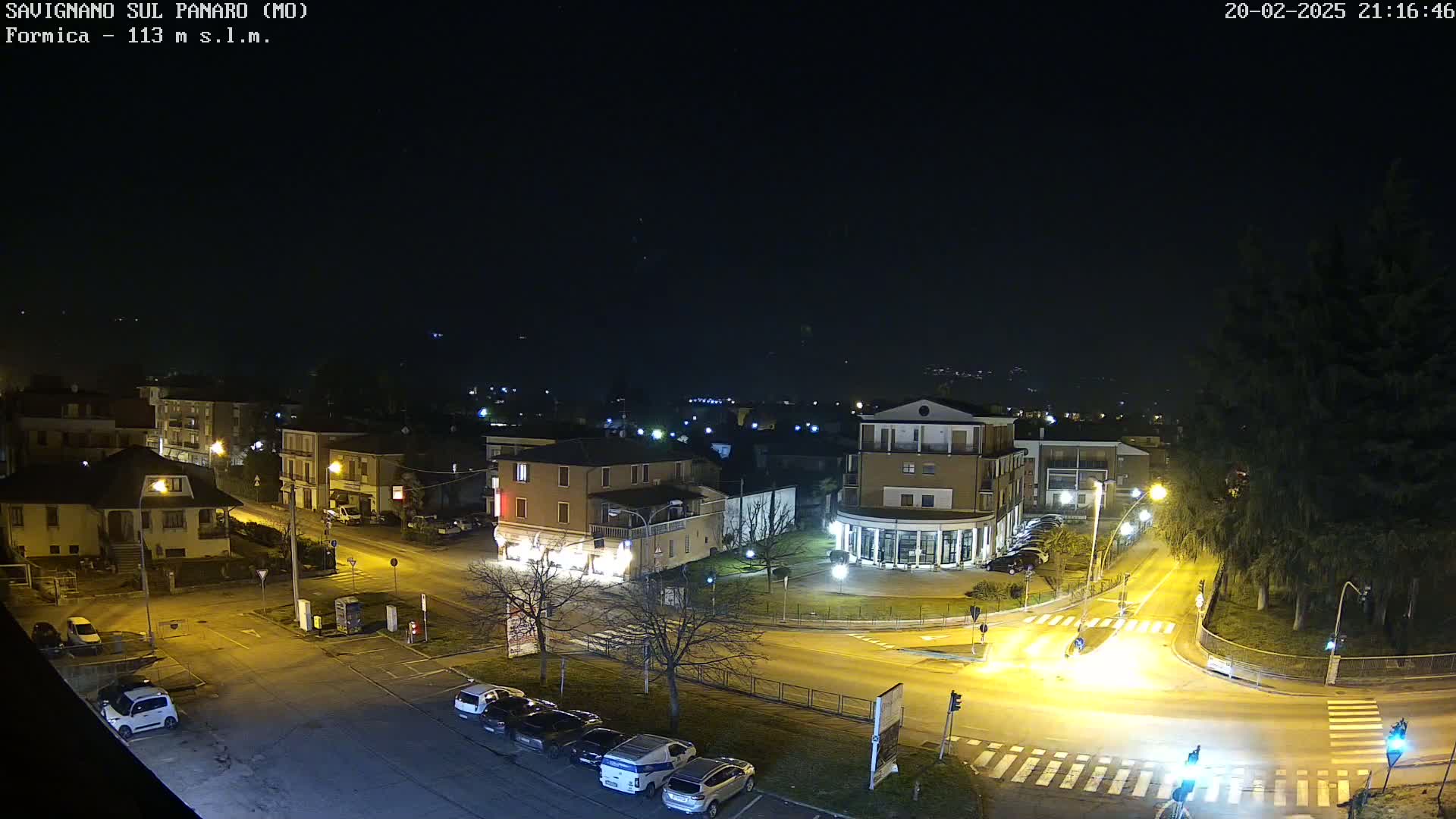 A nighttime aerial view of a town square with several buildings illuminated, cars parked in lots, and a few vehicles moving through a street intersection under a clear, dark sky.