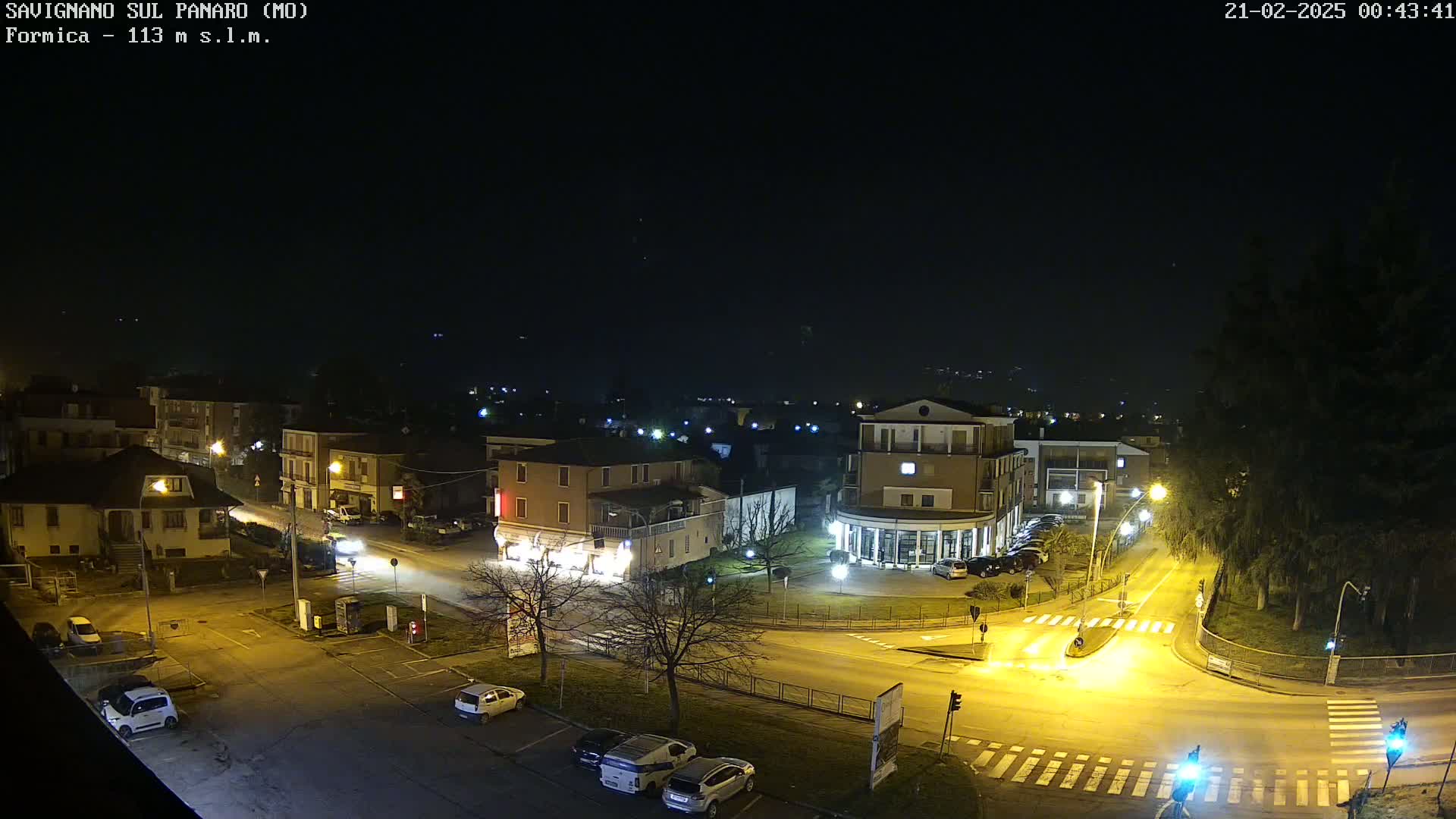 A nighttime, overhead view of a town square with cars parked and driving on illuminated roads, surrounded by buildings and trees under a clear, dark sky.