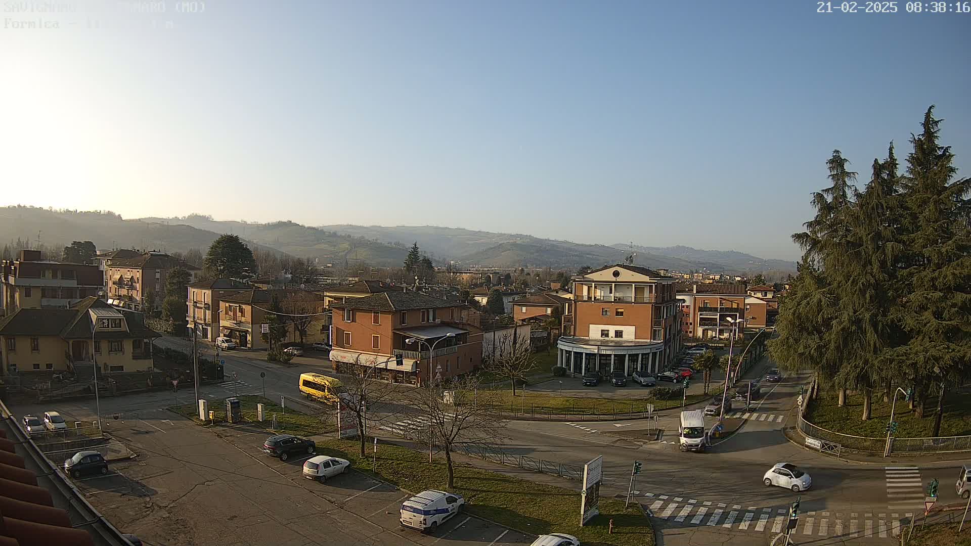 A sunny day reveals a town square with several buildings, cars, and a bus, set against a backdrop of rolling hills.