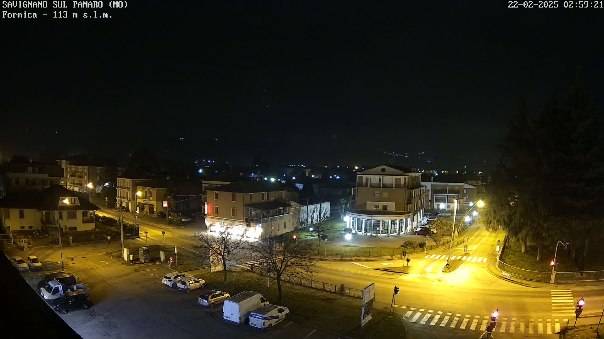 A nighttime aerial view of a town square with several buildings, cars parked along the streets, and streetlights illuminating the scene under a dark sky.