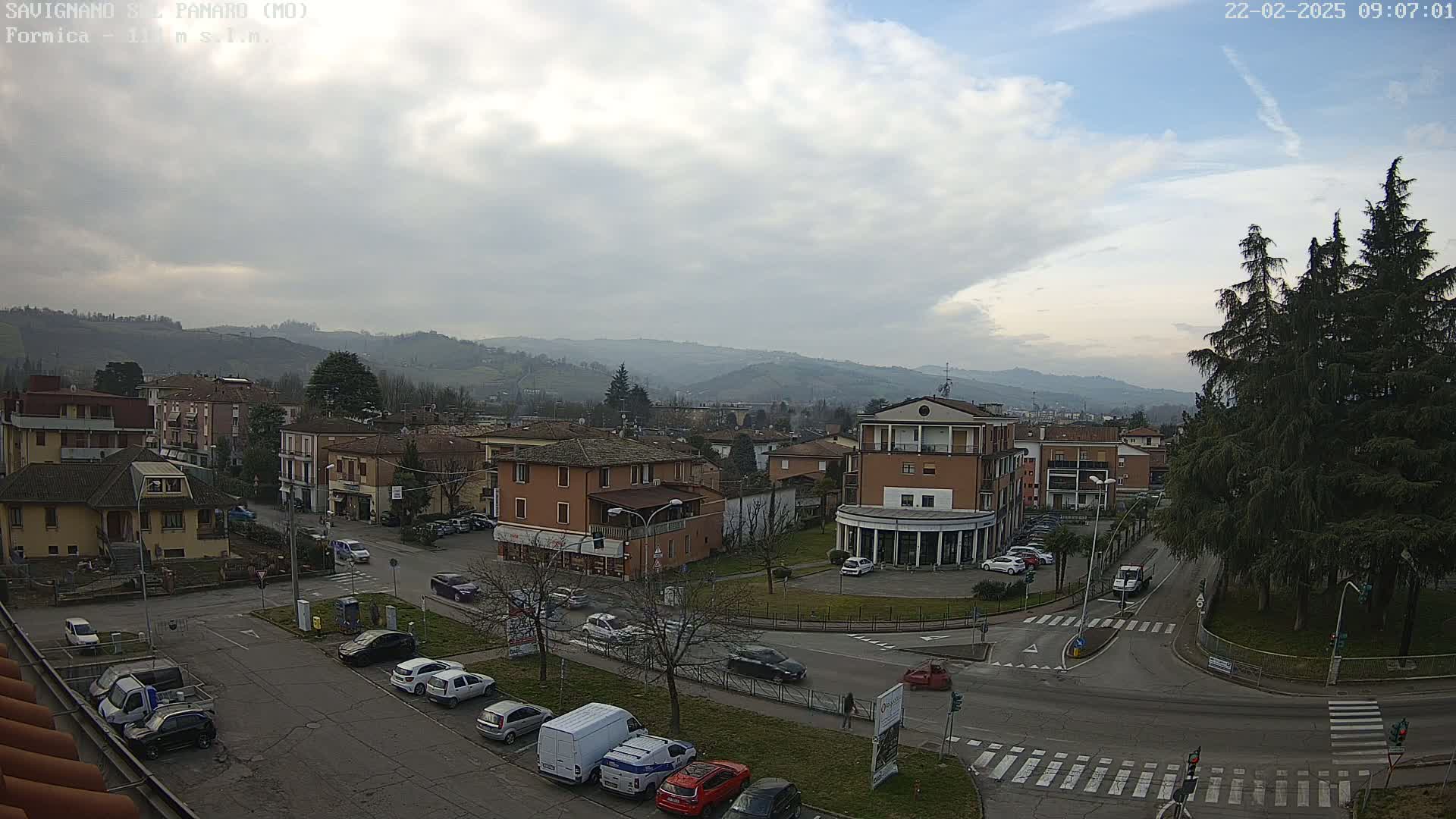 A mostly cloudy day reveals a small town nestled in a valley surrounded by hills, with cars parked and driving on the roads below.