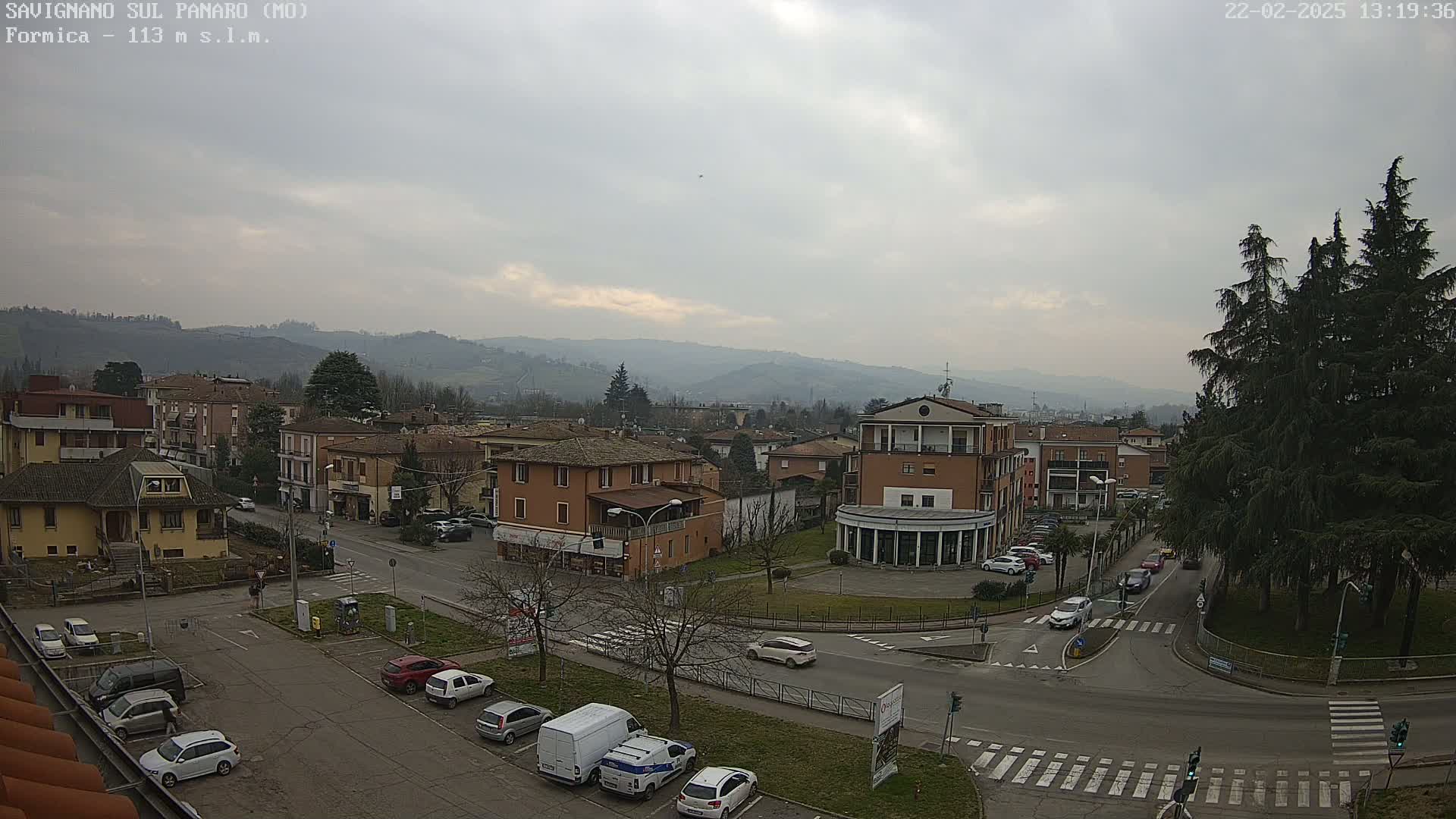 A mostly cloudy day reveals a view of a town nestled in a valley, with cars parked along the streets and several buildings visible, including a large building in the center.