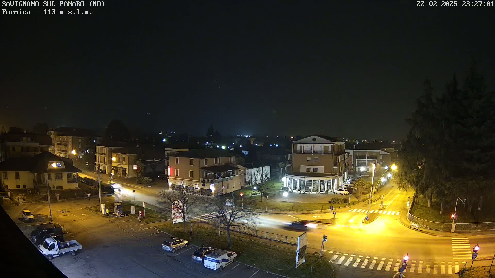 A nighttime aerial view of a town intersection with several cars and buildings illuminated by streetlights under a clear, dark sky.