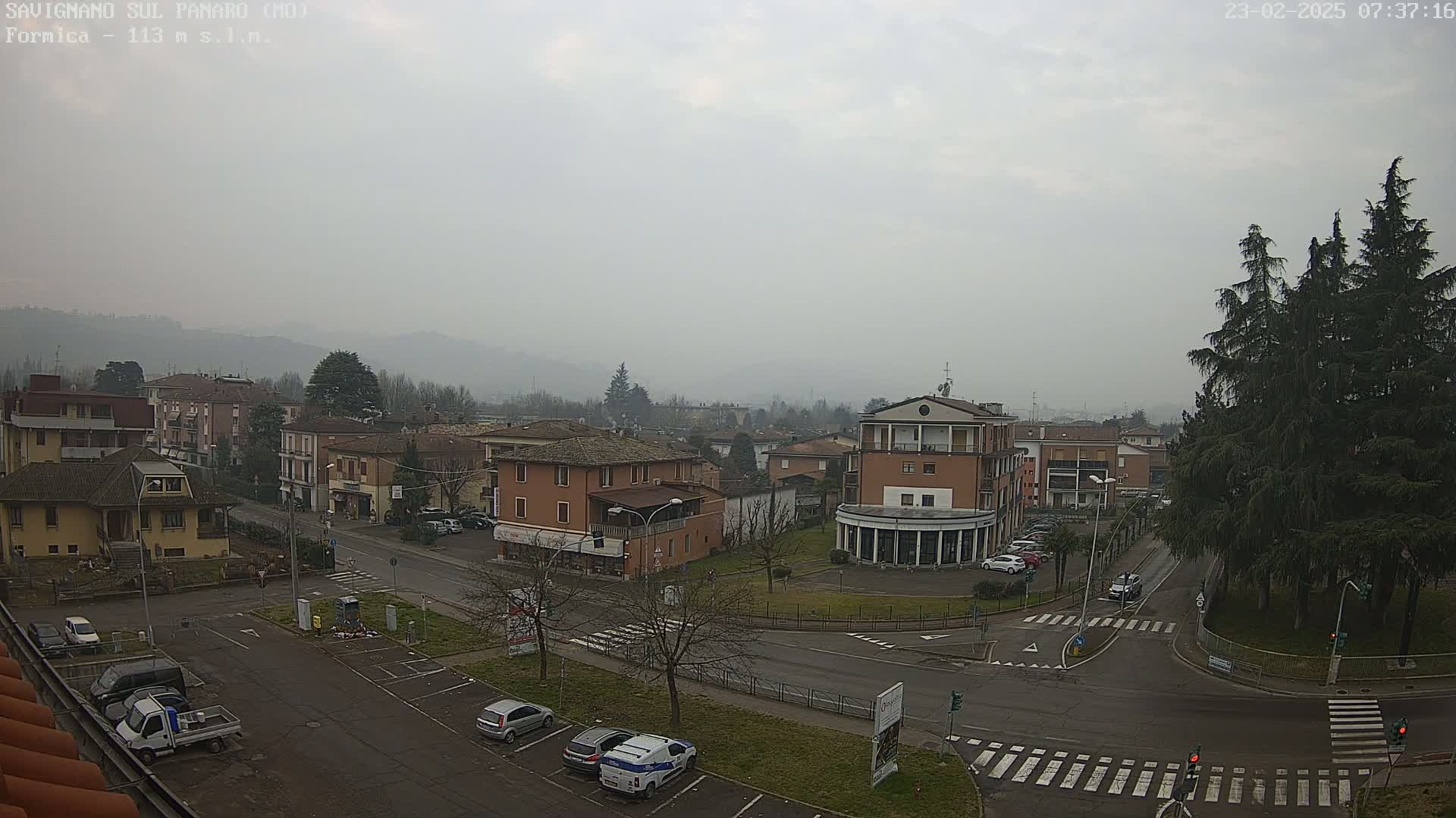 A mostly overcast view of a town square with several buildings, parked cars, and a few trees on a foggy day.