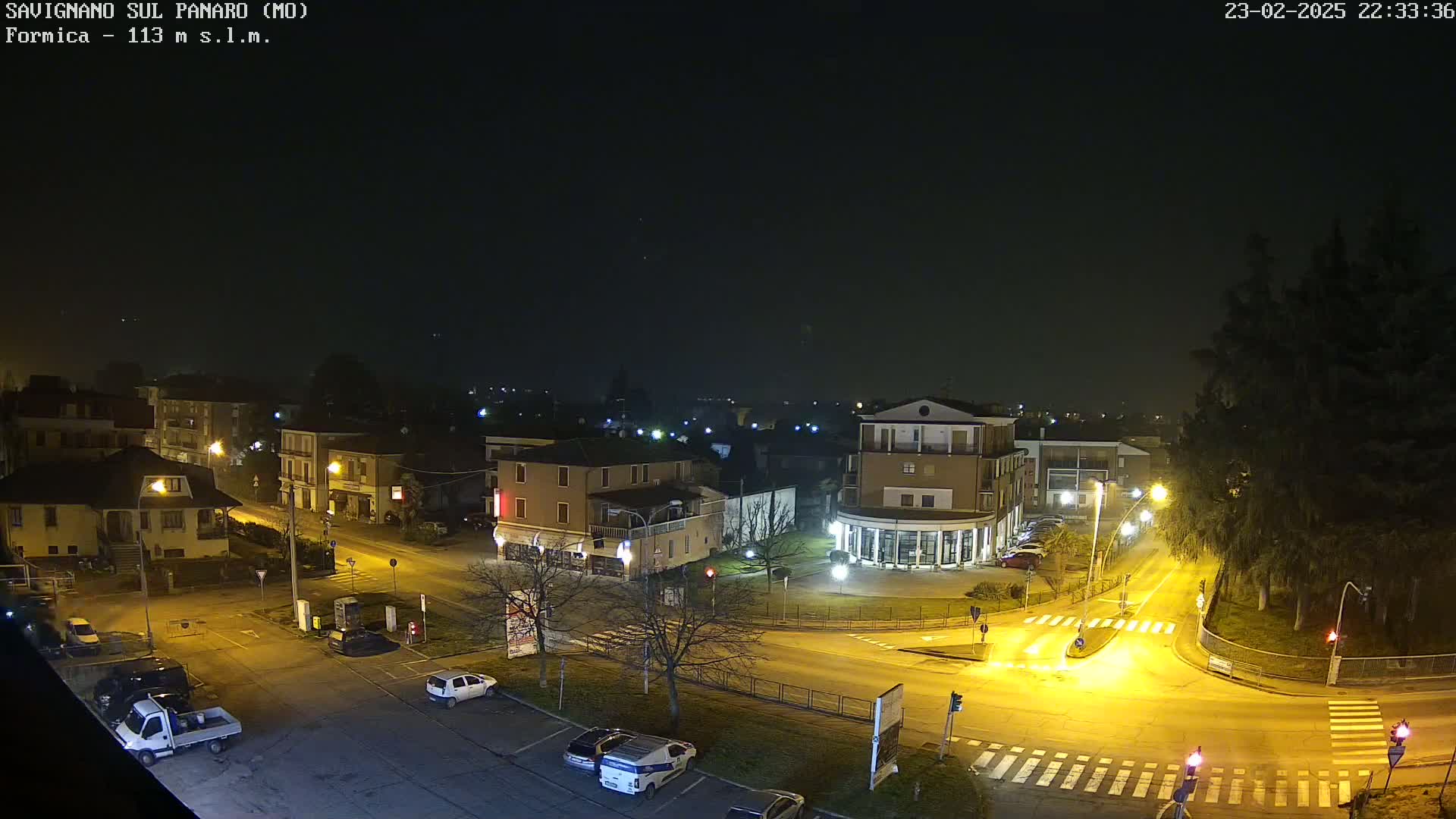 A nighttime aerial view of a town intersection with several cars and buildings illuminated by streetlights under a dark, slightly hazy sky.
