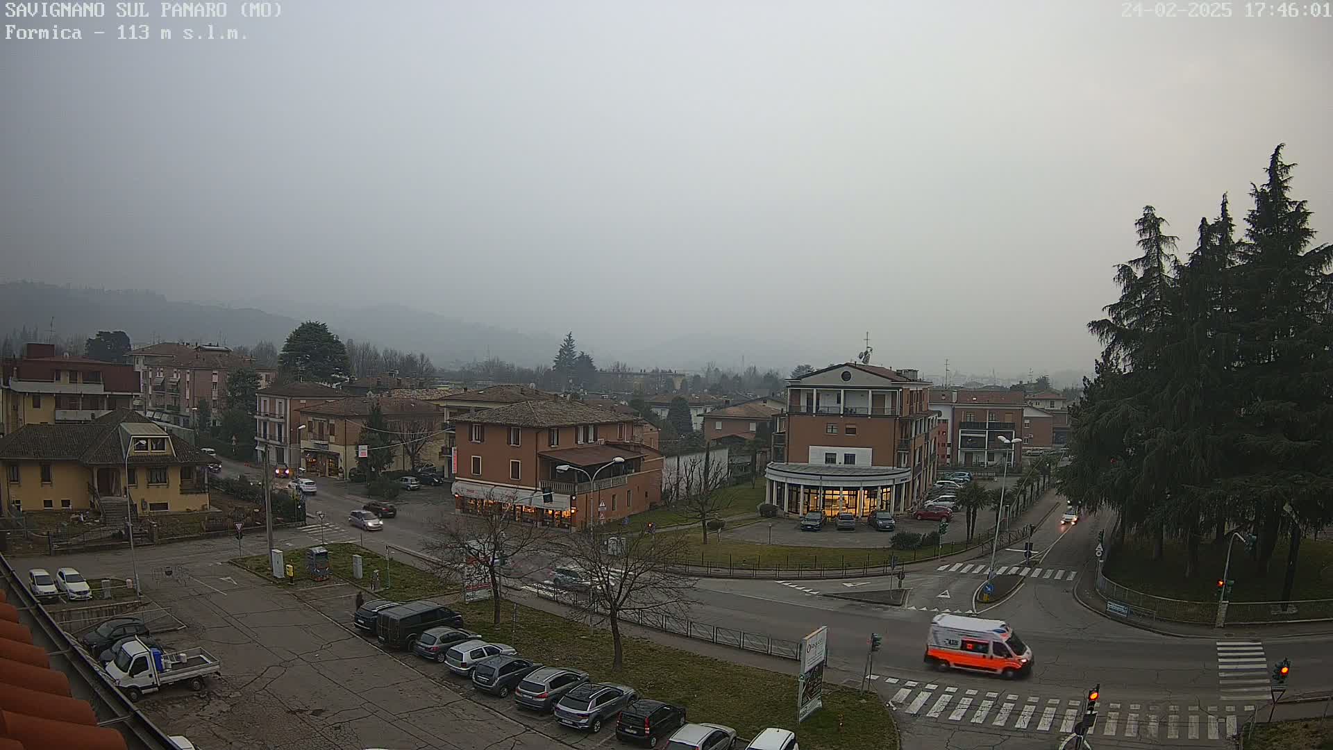 A hazy, overcast day reveals a town square with several buildings, parked cars, and an ambulance driving through an intersection.