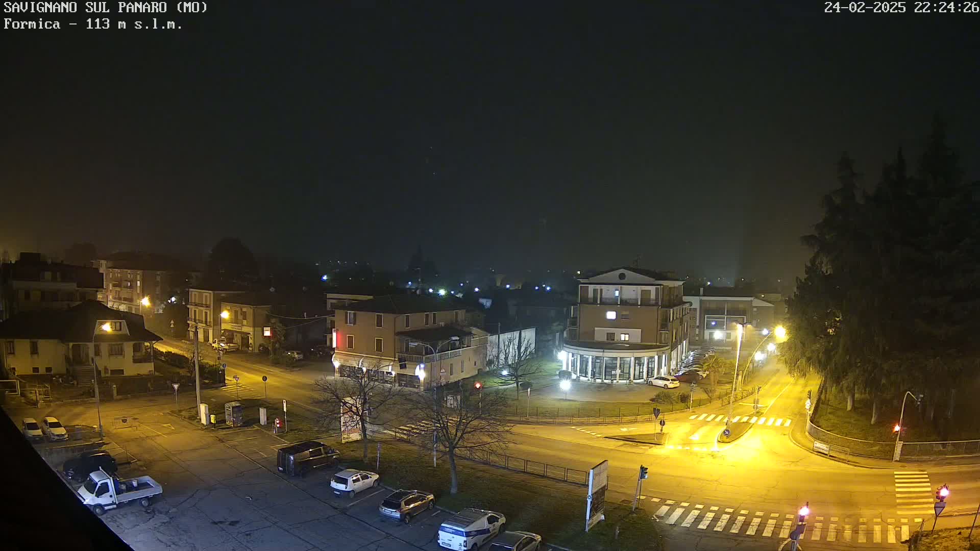 A nighttime, foggy view of a town square with cars parked in a lot and streetlights illuminating the roads and buildings.
