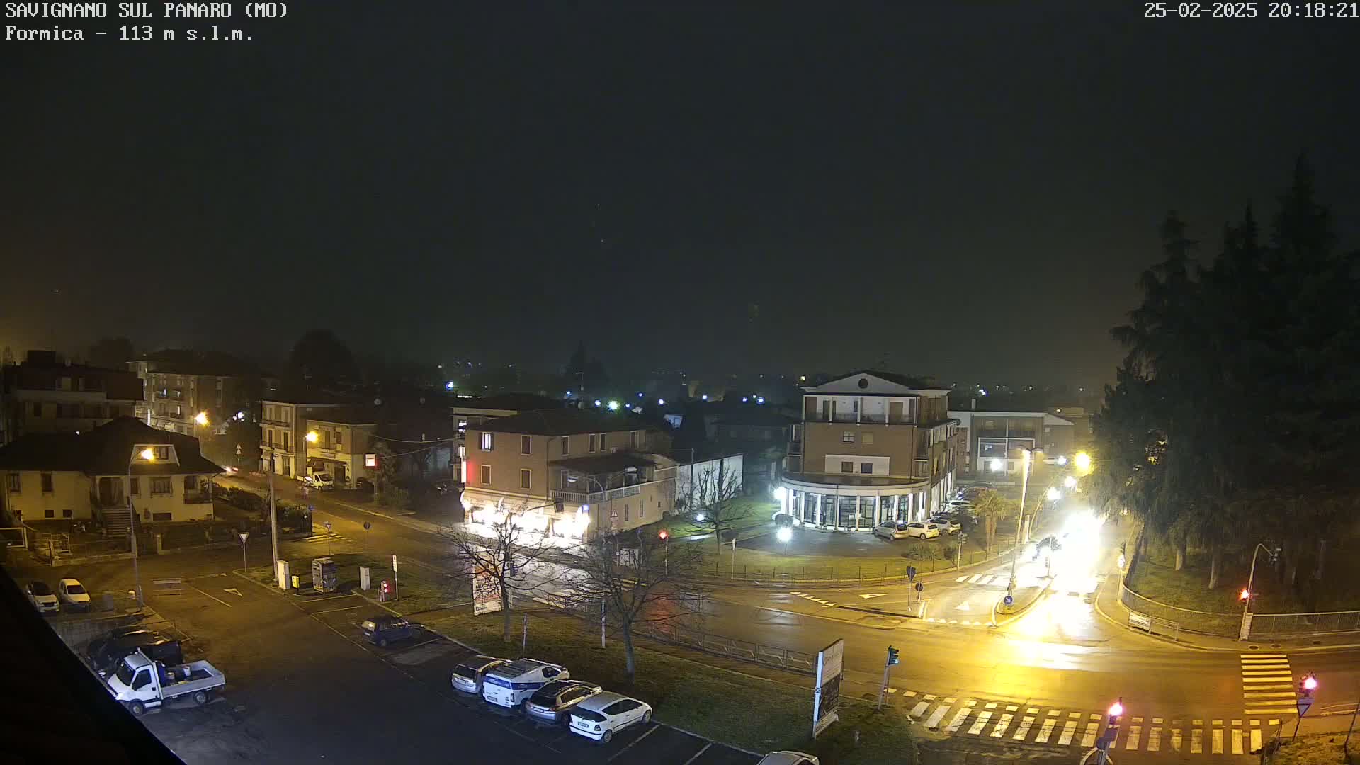 A nighttime view of a town square with wet roads and buildings under a foggy sky.