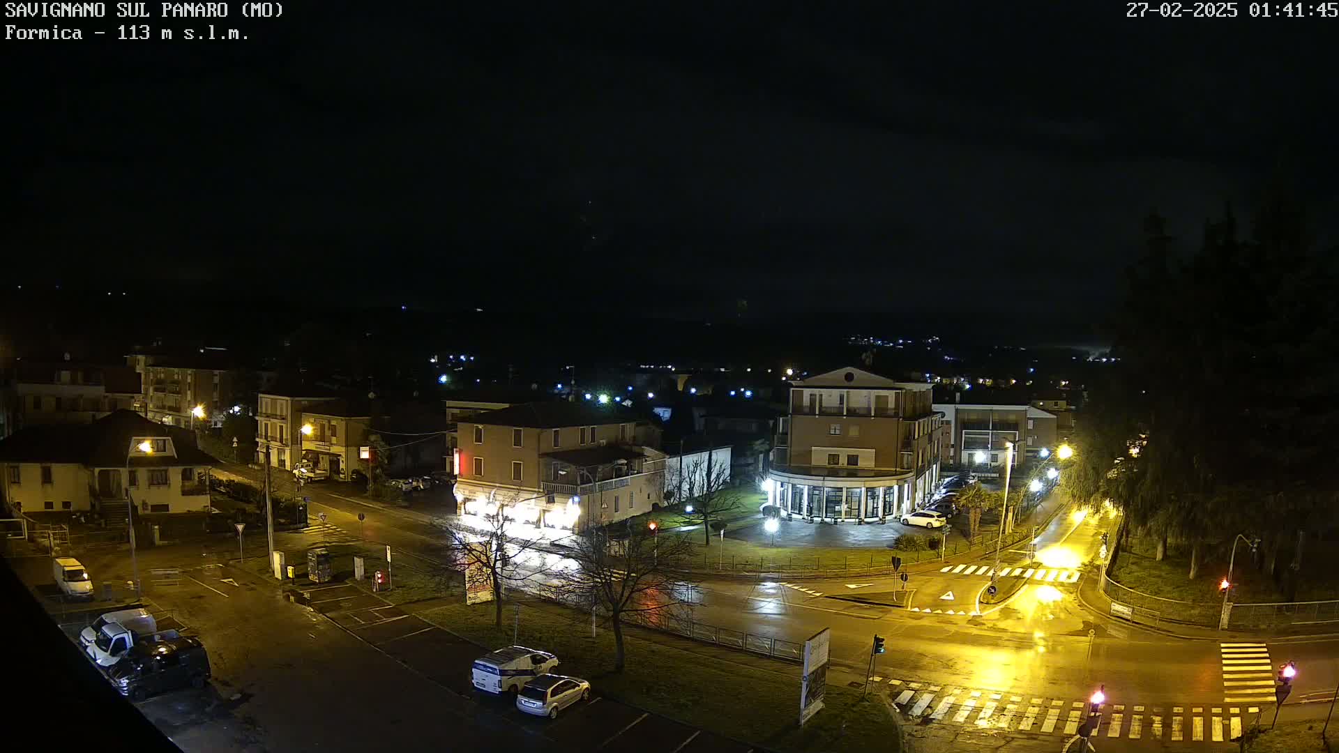 A nighttime view of a town's streets and buildings, wet from rain, with streetlights illuminating the wet roads and parked cars.