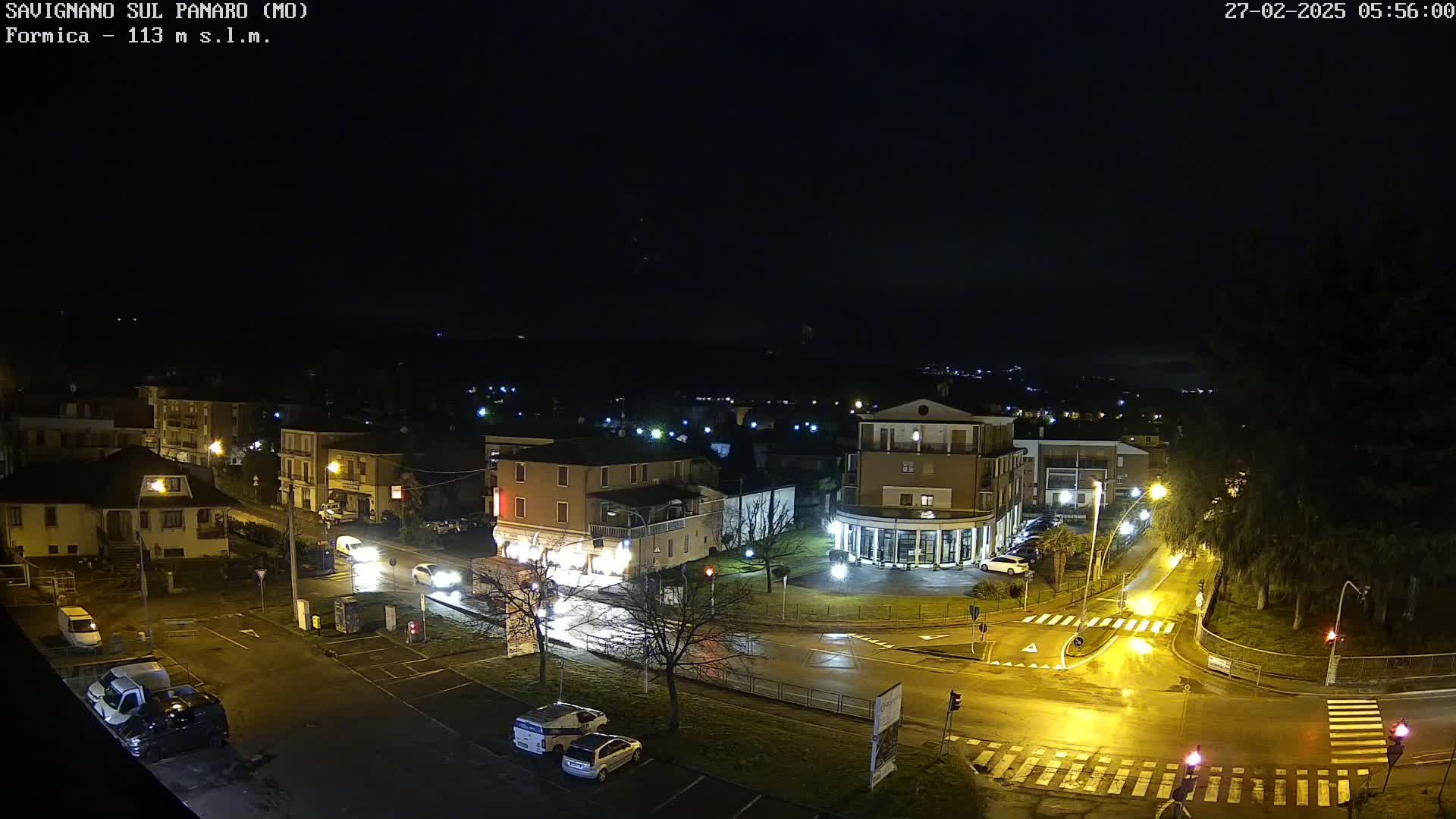 A wet, nighttime view of a town square with several cars parked and driving on roadways, lit by streetlights.